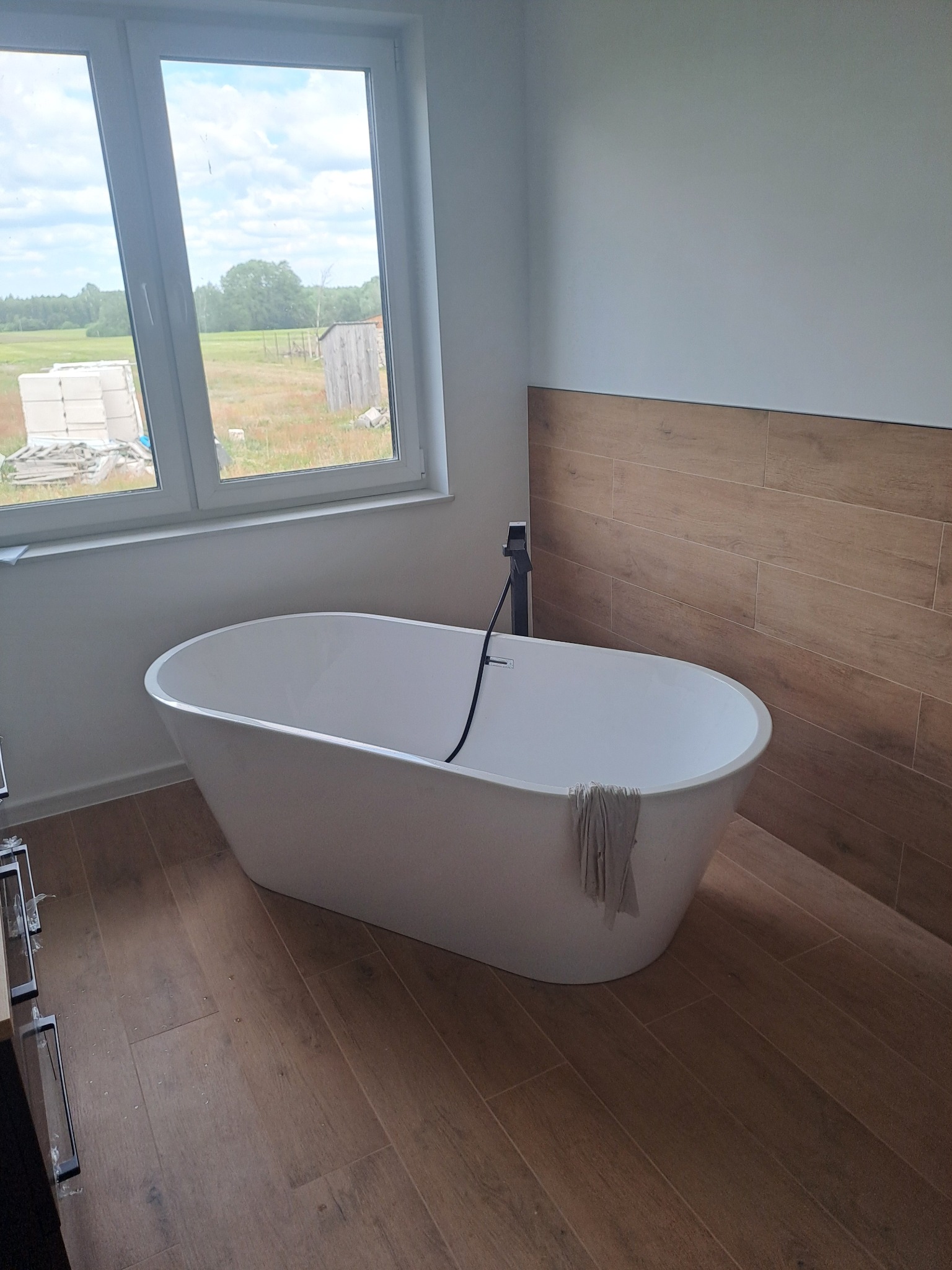 Wolnostojąca wanna w jasnej łazience z drewnopodobnymi płytkami na podłodze i części ściany, widok na wiejski krajobraz za oknem.