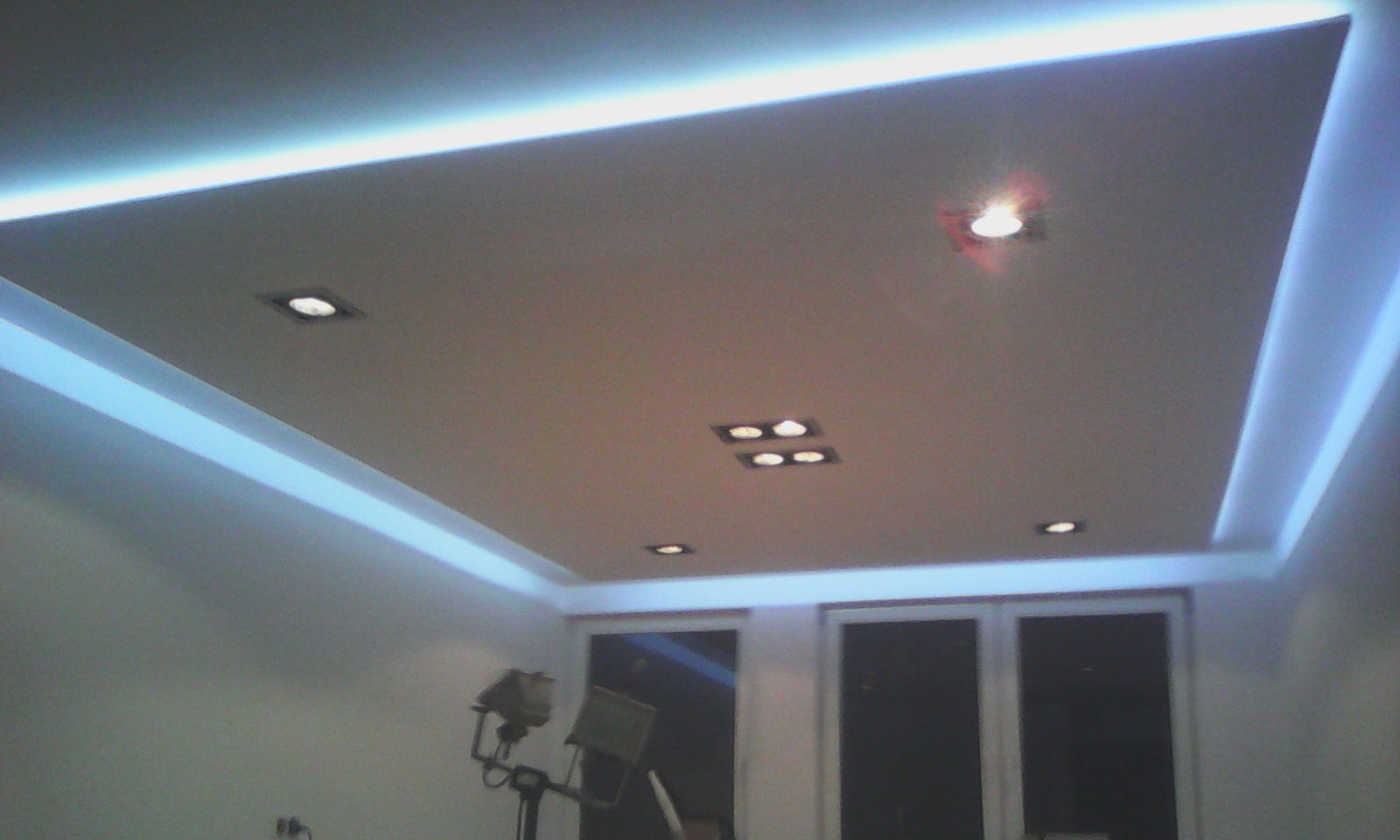 Podwieszany sufit z oświetleniem LED i punktowymi lampami w nowoczesnym pokoju, widoczny statyw oświetleniowy w rogu.
