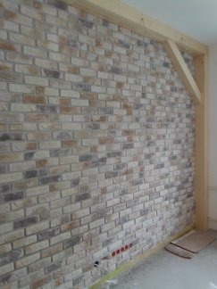 Ściana wykończona dekoracyjną cegłą w odcieniach beżu, szarości i brązu, z widoczną drewnianą konstrukcją ramy. Widoczne gniazdka elektryczne w dolnej części ściany.