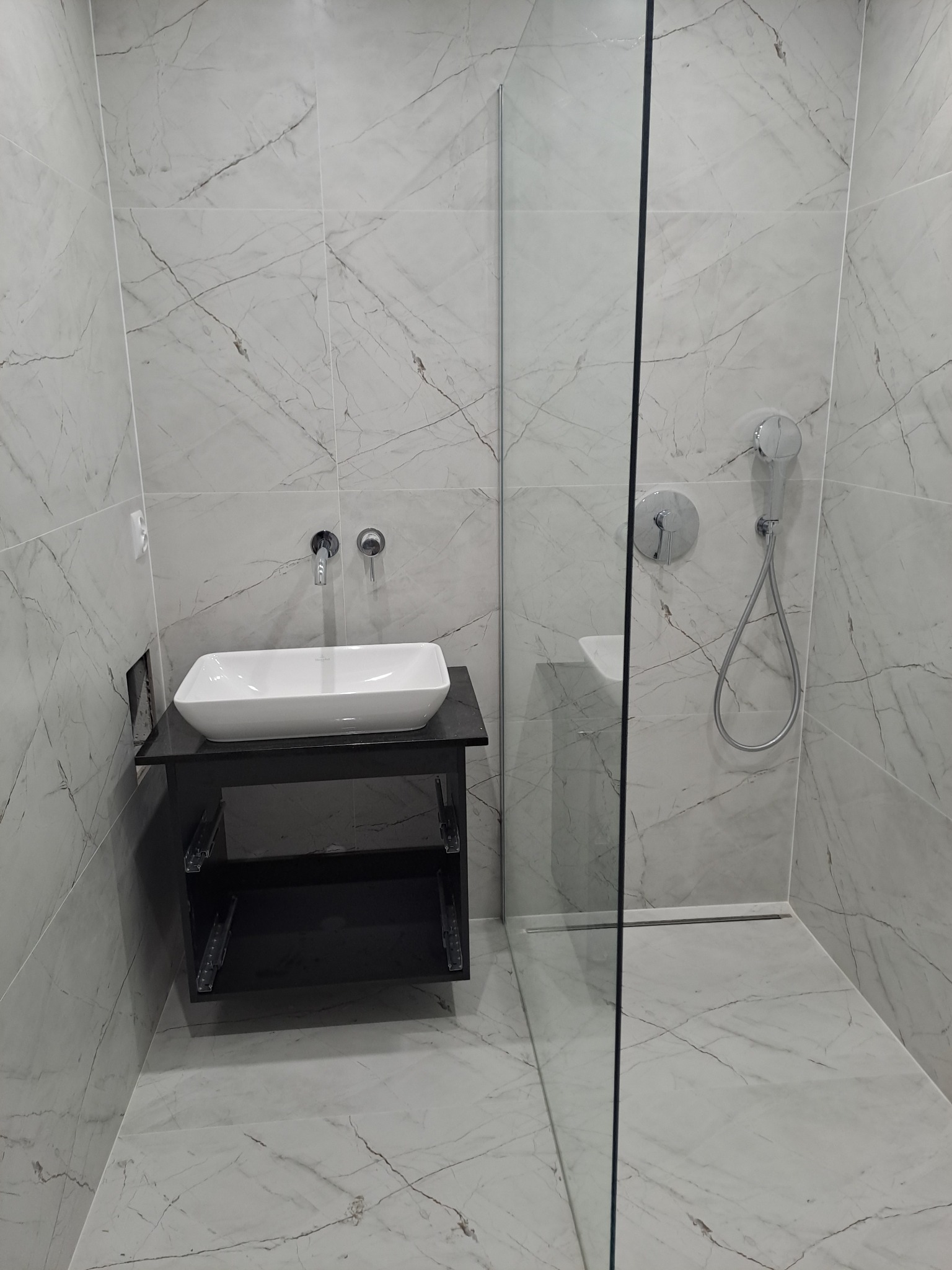 Nowoczesna łazienka z marmurowymi płytkami, kabiną prysznicową ze szkła i białą umywalką nablatową na czarnej szafce z otwartymi półkami, widoczny odpływ liniowy.