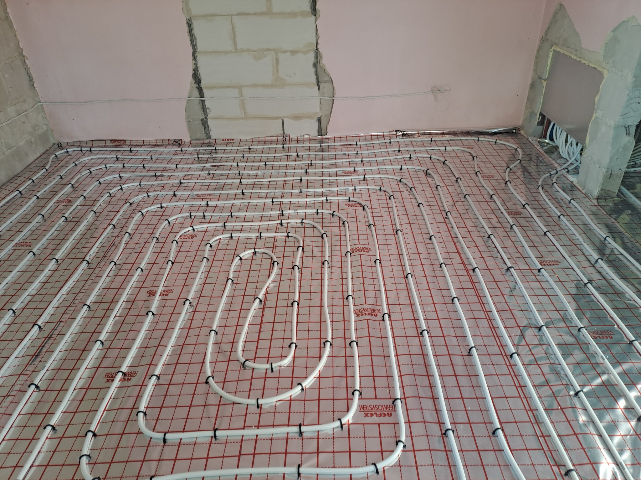 Białe rury ogrzewania podłogowego ułożone na czerwonej siatce w surowym wnętrzu łazienki przed wylaniem posadzki.