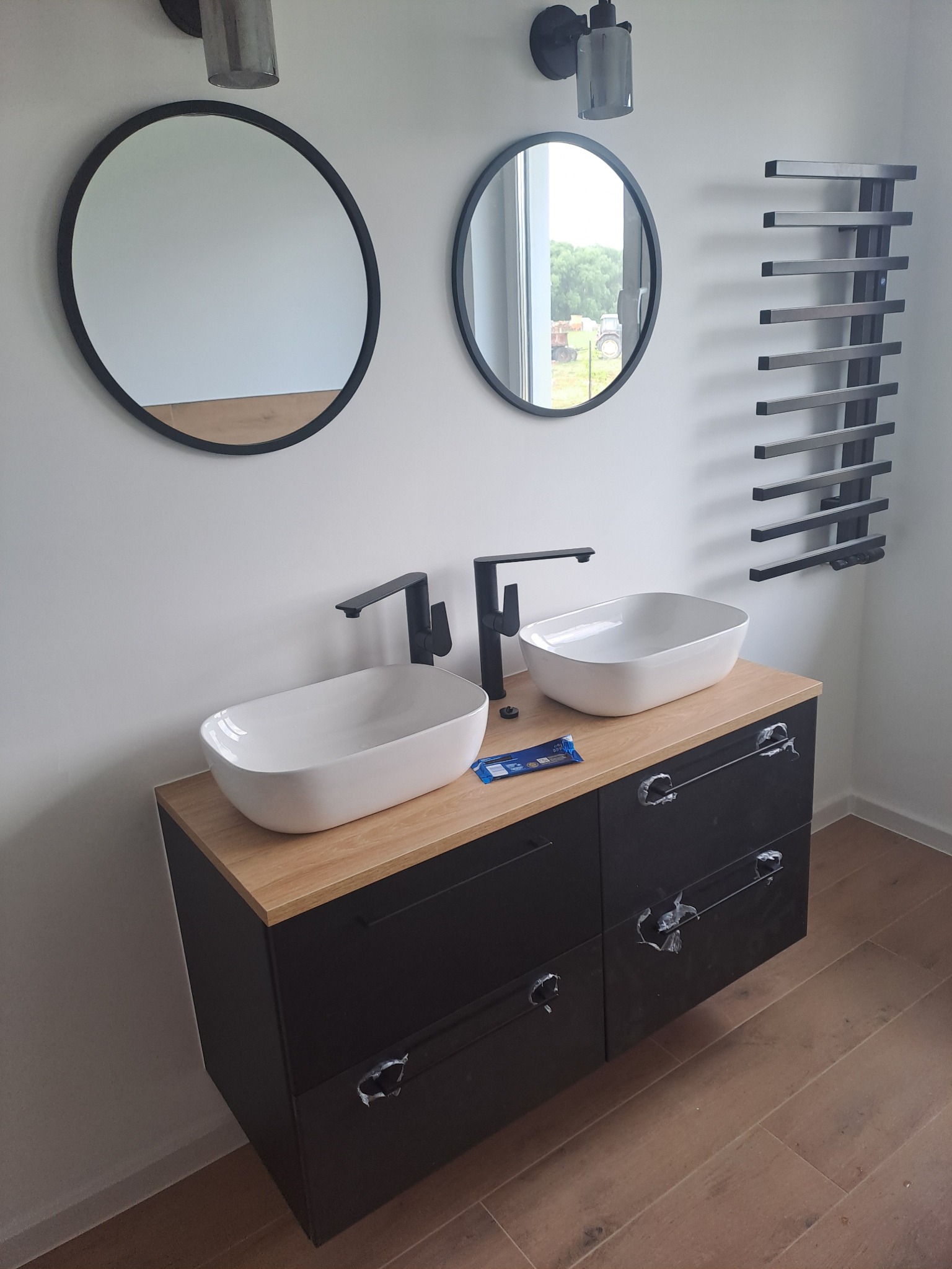 Nowoczesna łazienka z dwoma okrągłymi lustrami, dwiema owalnymi umywalkami nablatowymi, czarną szafką z drewnianym blatem i grzejnikiem drabinkowym w kolorze czarnym. Na blacie leżą niebieskie...