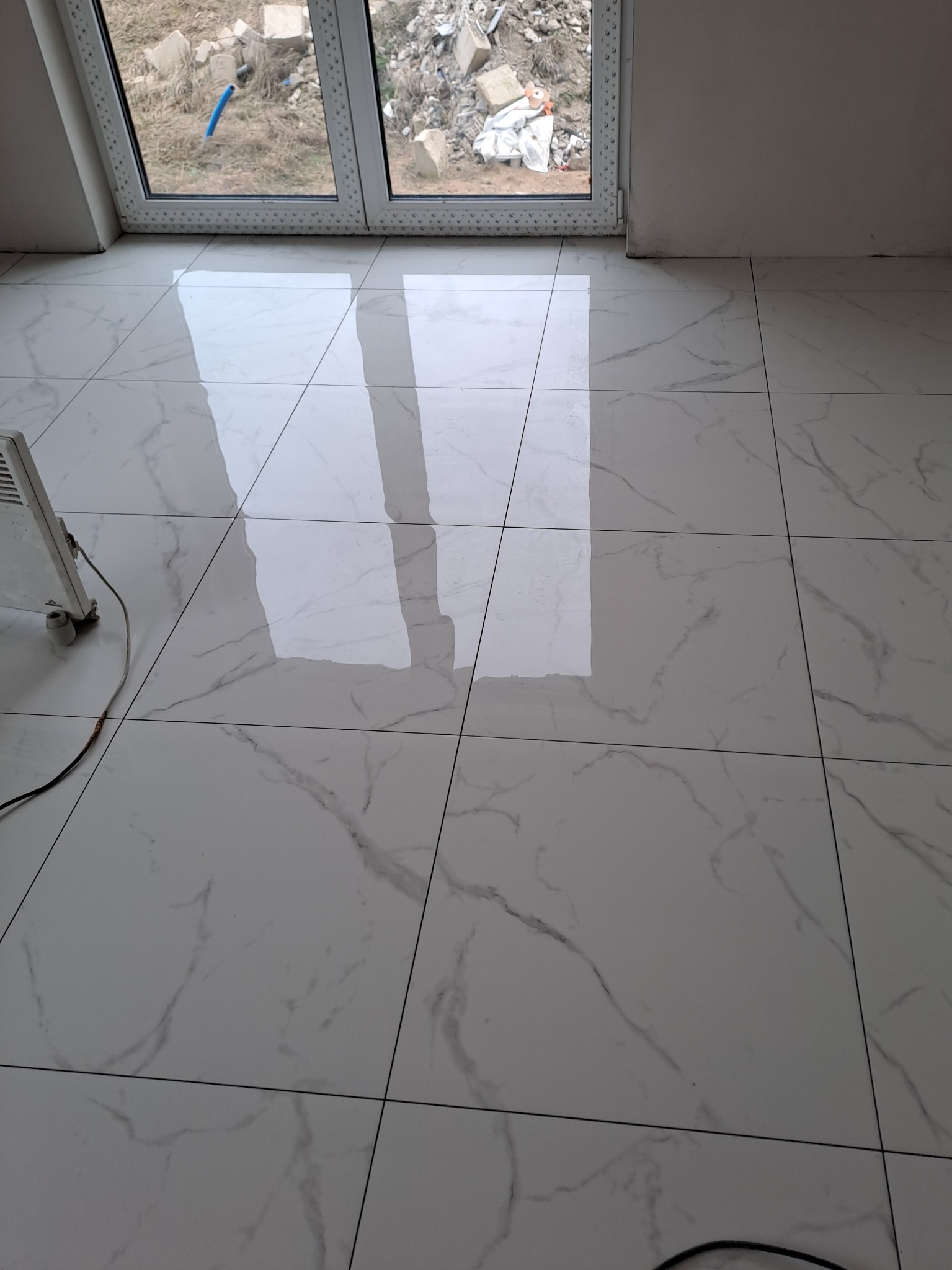 Błyszcząca, biała podłoga z płytek imitujących marmur, świeżo po ułożeniu, widoczny grzejnik elektryczny i okno z widokiem na gruzowisko.