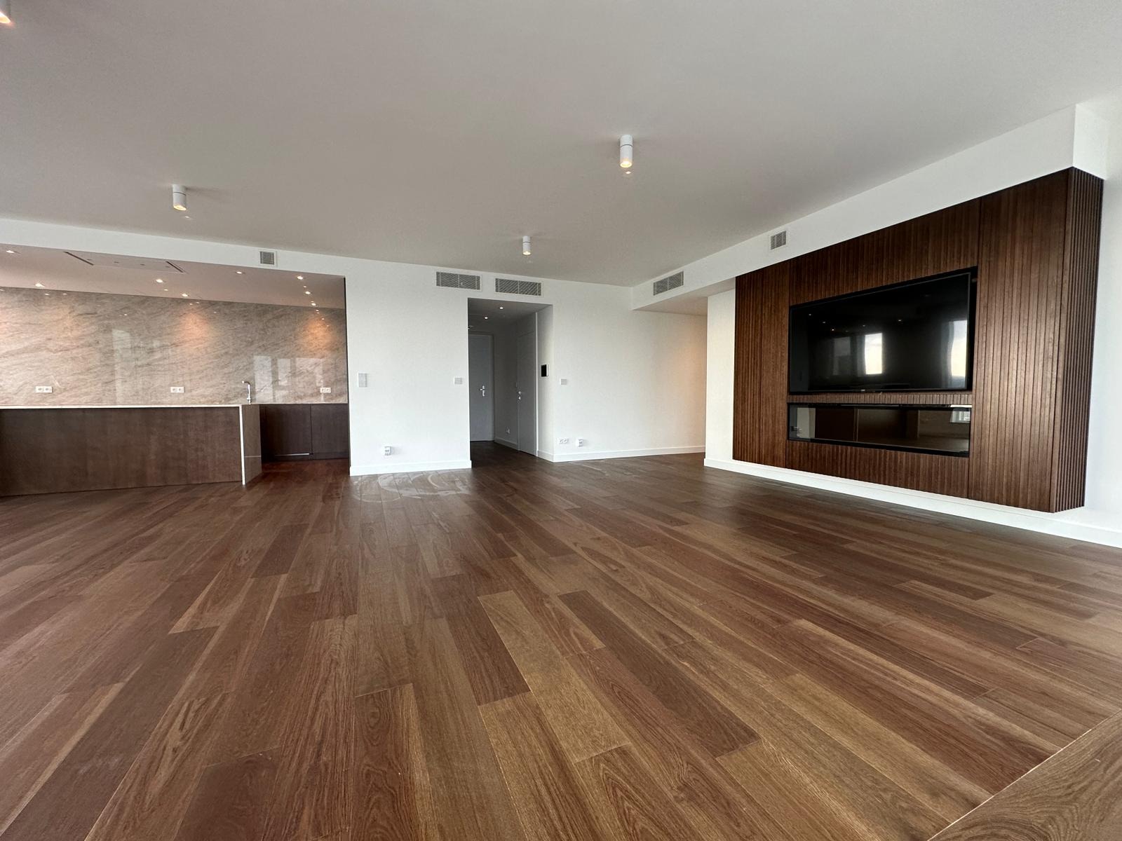 Przestronne, jasne wnętrze apartamentu z drewnianą podłogą, ścianą z telewizorem obudowaną w ciemne drewno oraz widoczną w tle zabudową kuchenną z marmurowym blatem.