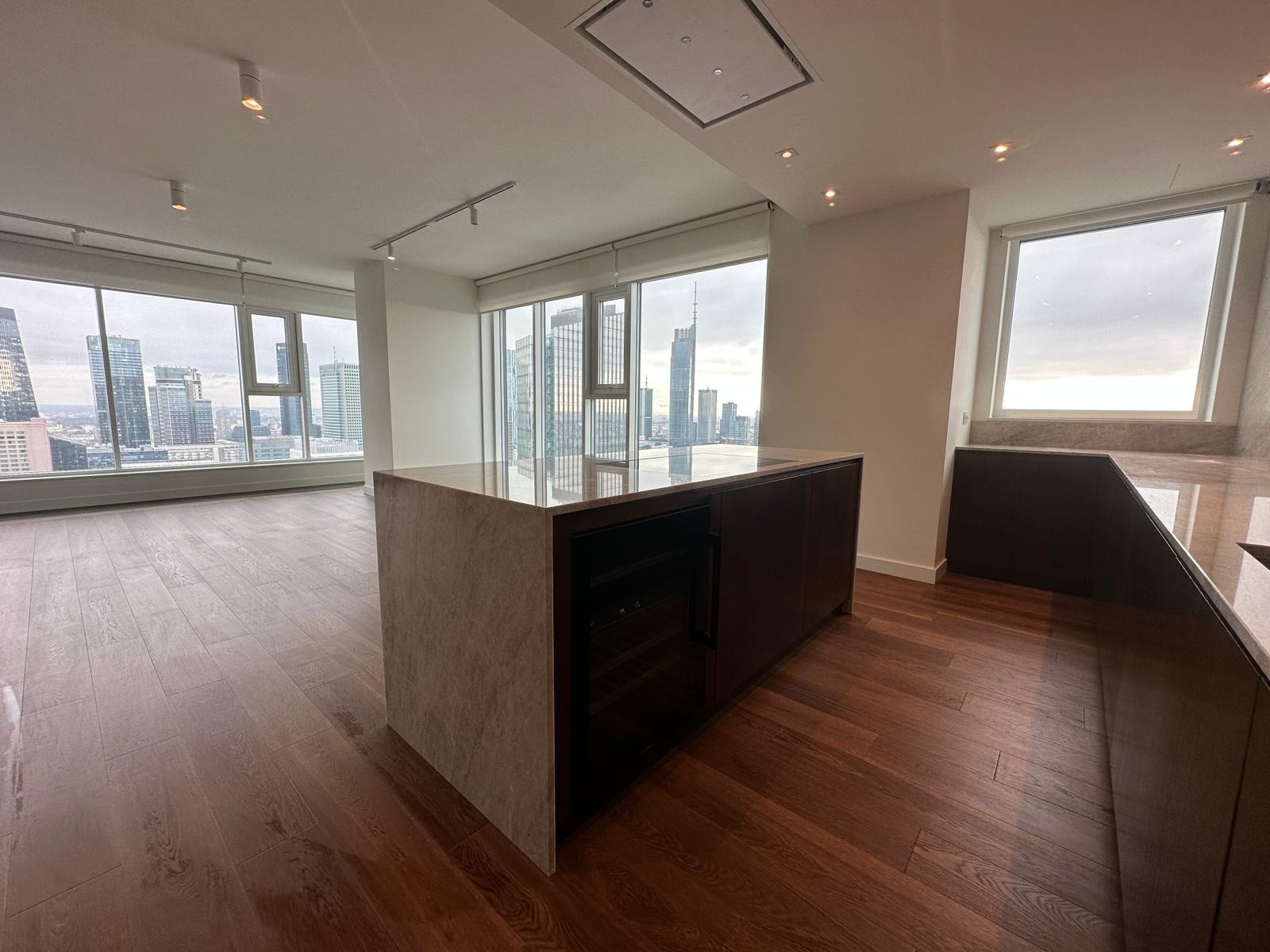 Nowoczesna wyspa kuchenna z wbudowaną chłodziarką do wina, drewniana podłoga, widok na panoramę miasta przez duże okna.