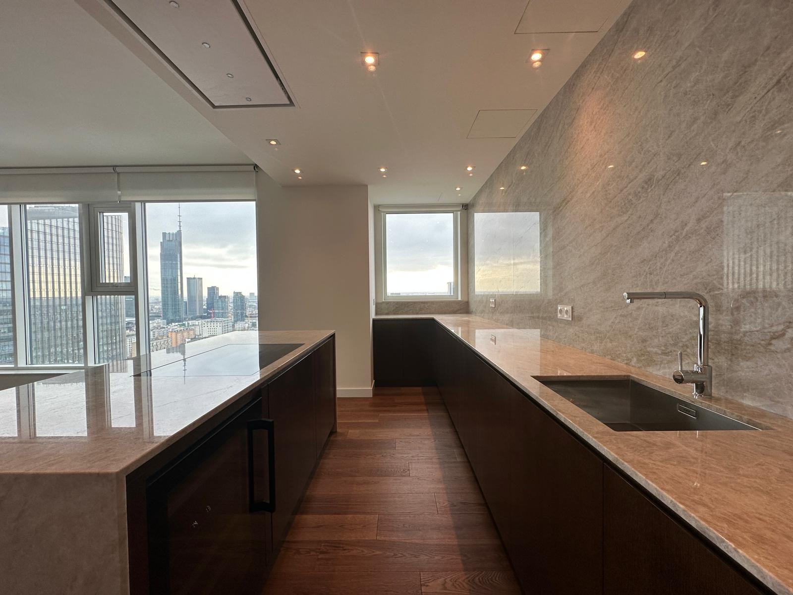 Nowoczesna kuchnia z marmurowymi blatami i ścianą, widok na panoramę miasta przez duże okna, drewniana podłoga, minimalistyczne oświetlenie sufitowe.