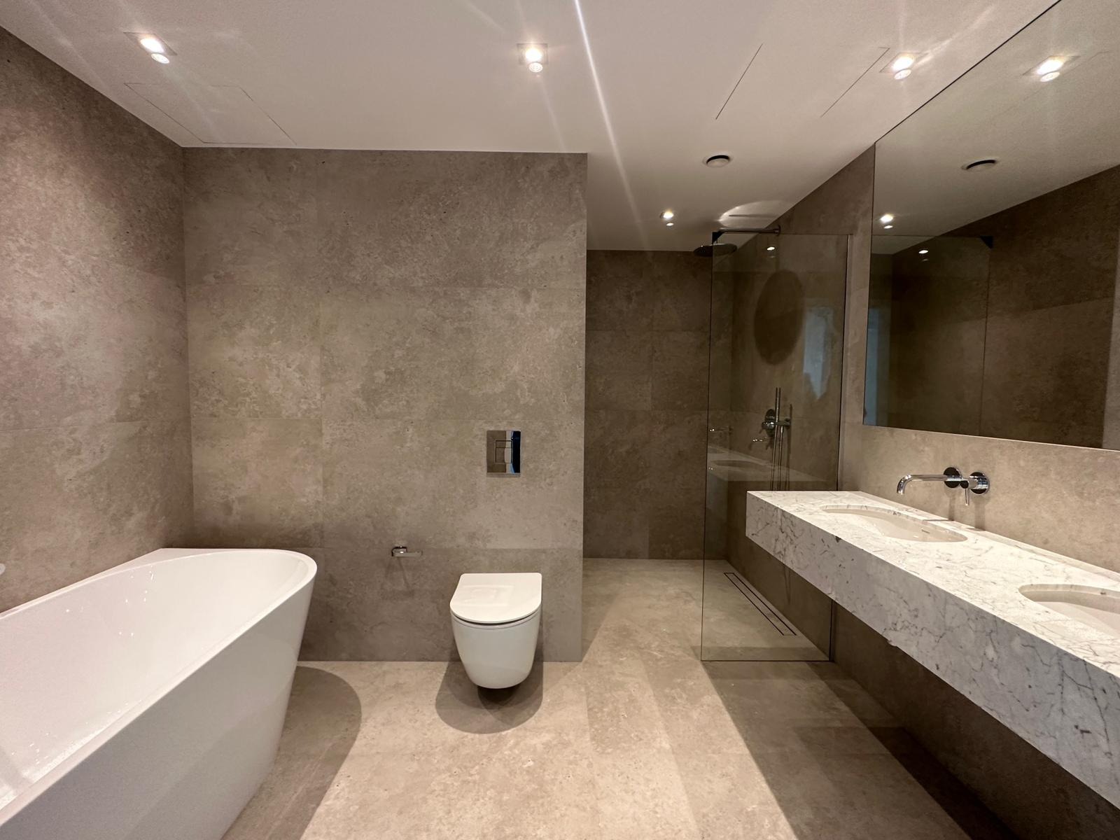 Nowoczesna łazienka z beżowymi płytkami, wolnostojącą białą wanną, podwójną umywalką na marmurowym blacie i kabiną prysznicową ze szklaną ścianą.