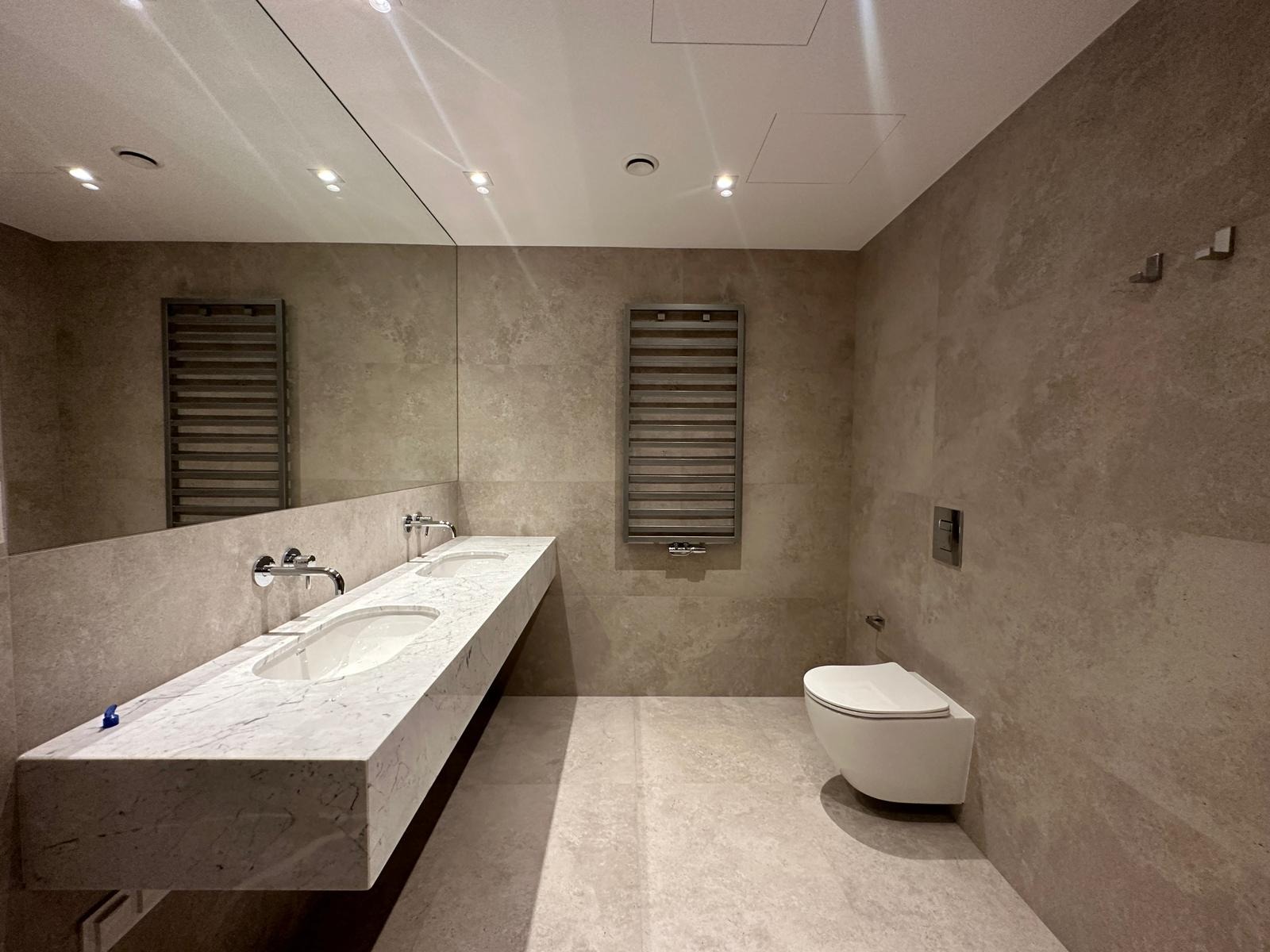 Nowoczesna łazienka w stonowanych beżach z długim, marmurowym blatem z dwiema umywalkami, podwieszaną toaletą i grzejnikiem drabinkowym, oświetlona punktowymi lampami sufitowymi.
