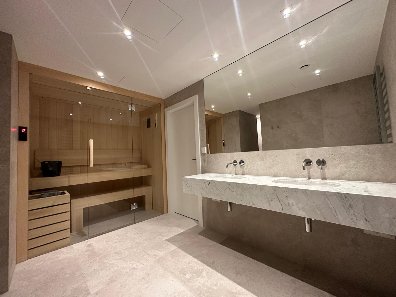 Nowoczesna łazienka z sauną: drewniana sauna za szklaną ścianą, marmurowa umywalka z dwoma kranami, szara ściana i podłoga, oświetlenie punktowe w suficie.