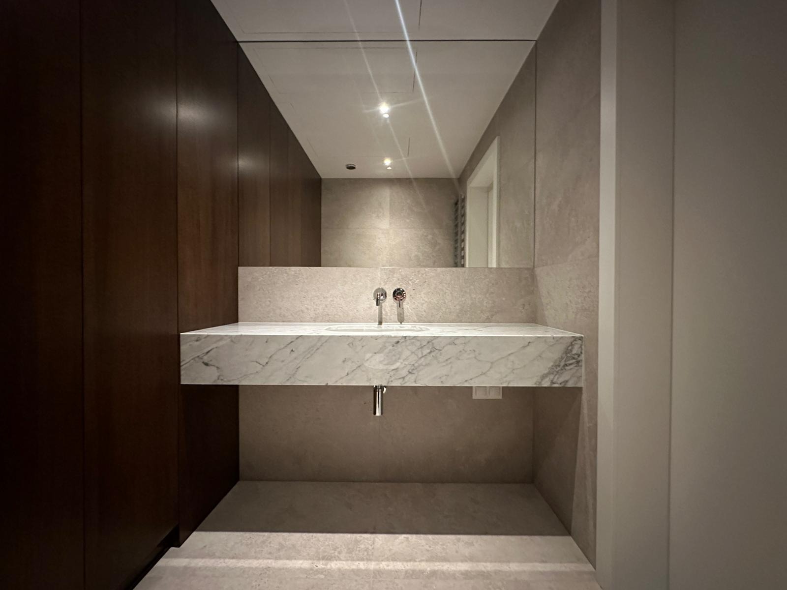 Nowoczesna łazienka z marmurową umywalką wiszącą, minimalistyczną armaturą i dużym lustrem bez ramy, z widocznymi elementami zabudowy stolarskiej w ciemnym drewnie.