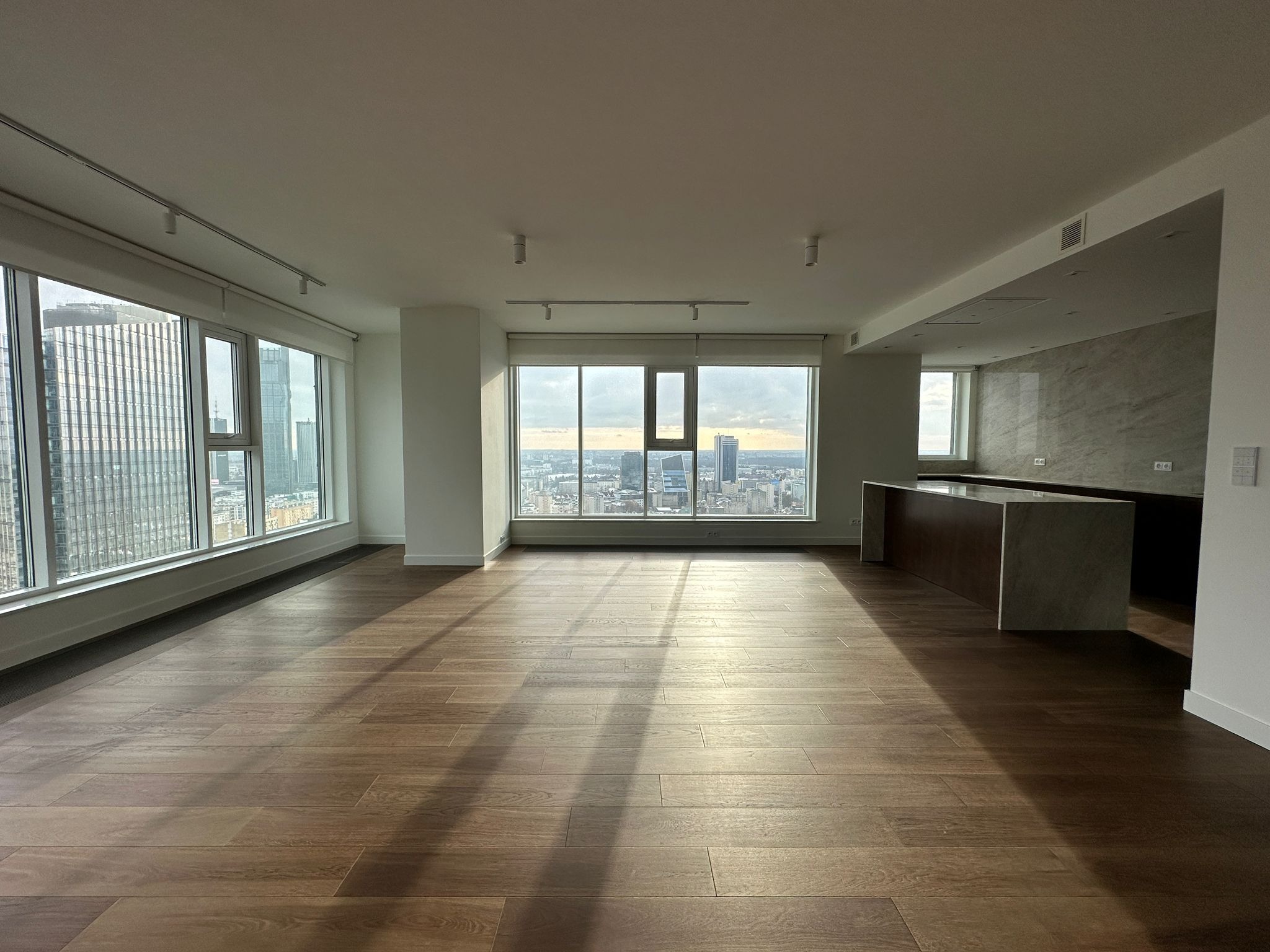 Wnętrze apartamentu z parkietem w odcieniach brązu, widok na panoramę miasta przez duże okna, oświetlenie szynowe i częściowo widoczna zabudowa kuchenna z blatem z imitacji kamienia.