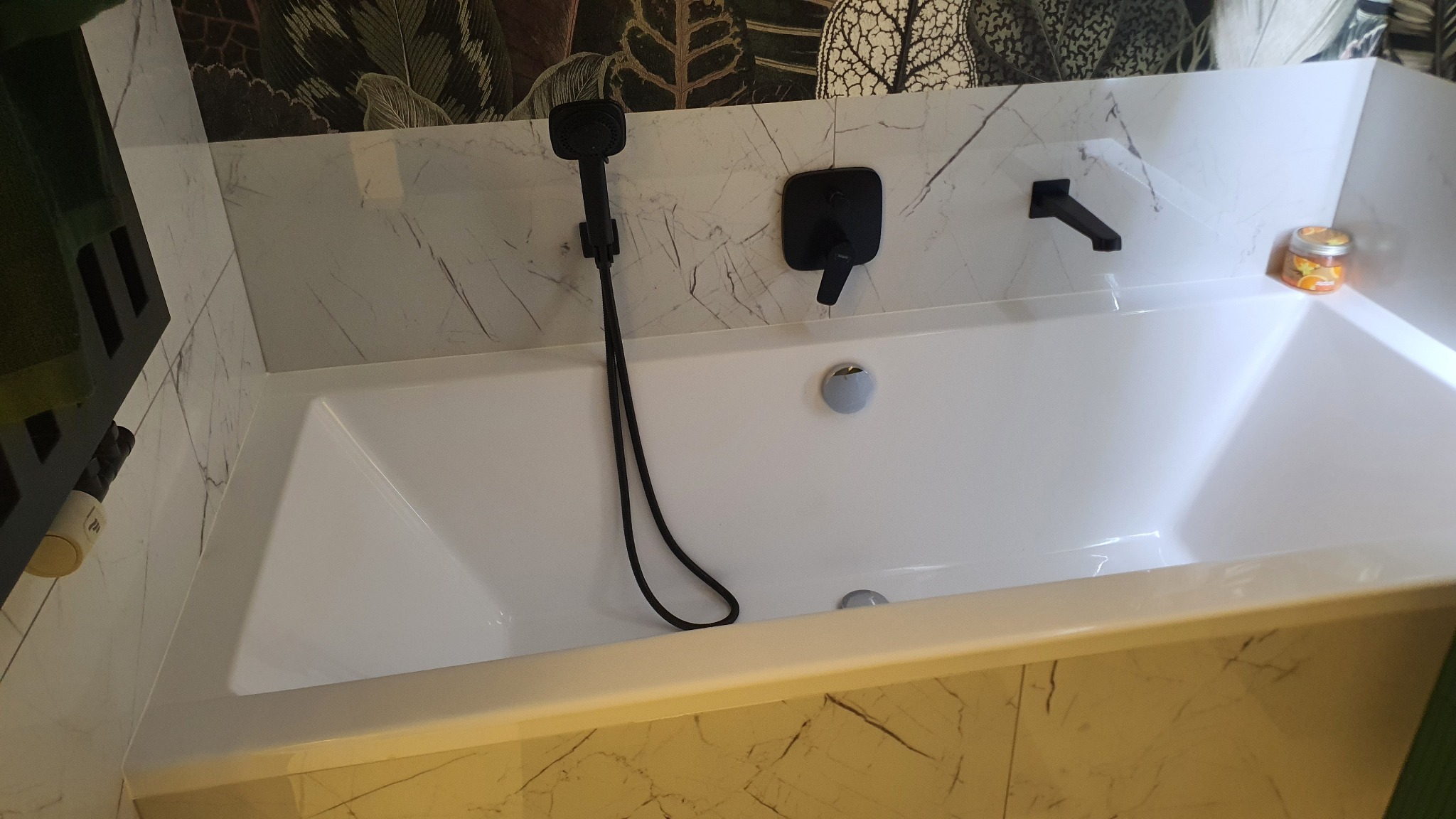 Biała wanna z czarną armaturą, marmurowe płytki na ścianach i egzotyczny wzór tapety w tle. Na wannie stoi słoik z pomarańczowymi plasterkami.