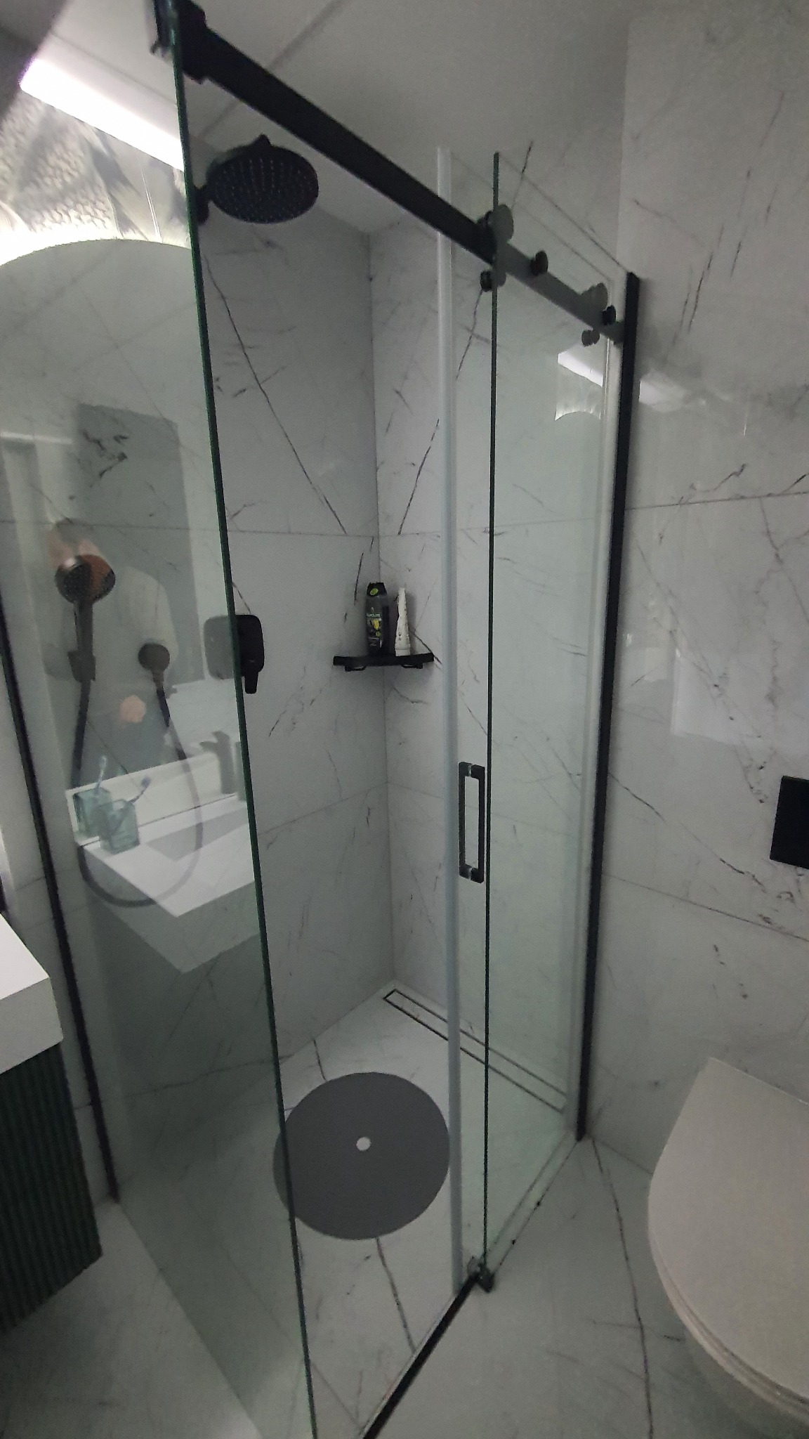 Nowoczesna kabina prysznicowa z czarnymi detalami, marmurowe płytki na ścianach, okrągły odpływ liniowy w podłodze, widoczna armatura i półka z kosmetykami.