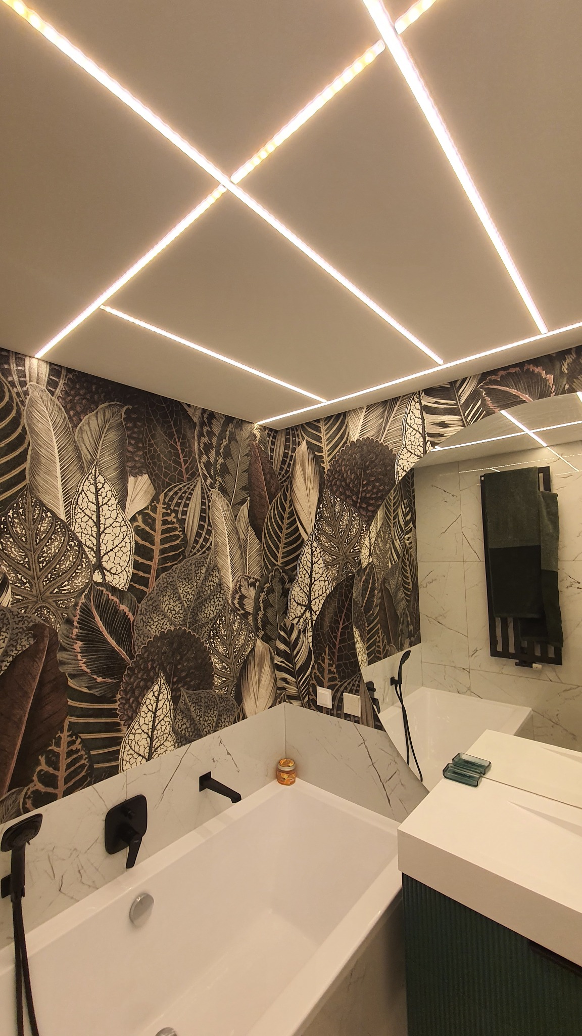 Nowoczesna łazienka z wanną obudowaną marmurem, czarną armaturą i ścianą w tapetę z motywem liści, oświetlona ledowymi listwami na suficie.