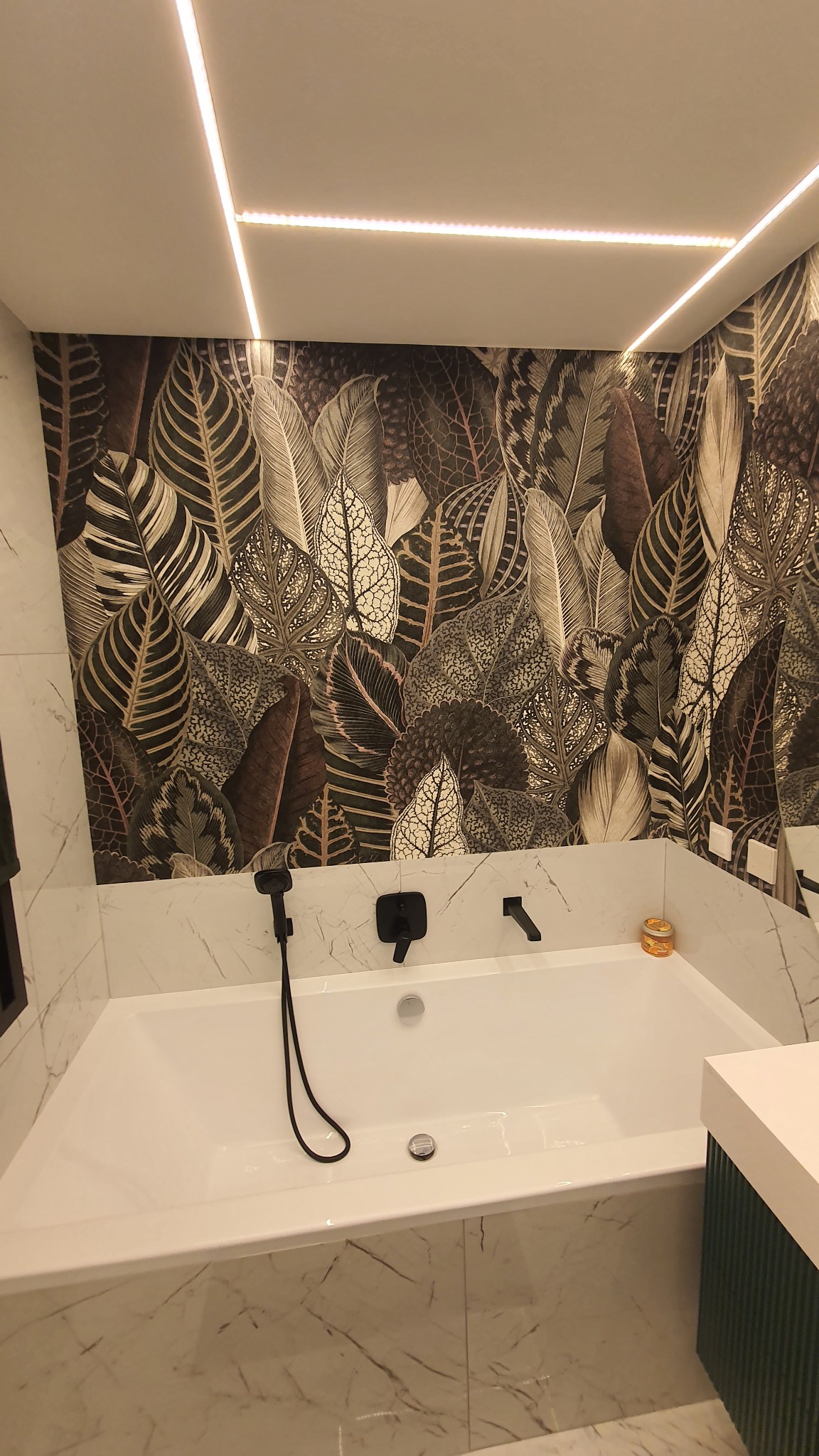 Nowoczesna łazienka z białą wanną obudowaną marmurem, czarna armatura i tapeta w motywy liści, oświetlenie LED w suficie.