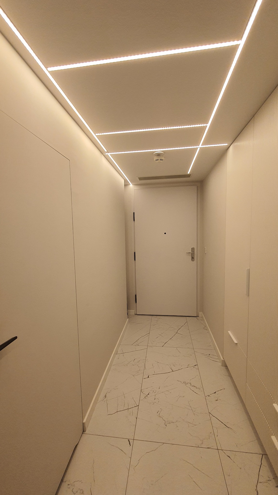 Nowoczesny korytarz z białymi ścianami, marmurową podłogą i geometrycznym oświetleniem LED na suficie.