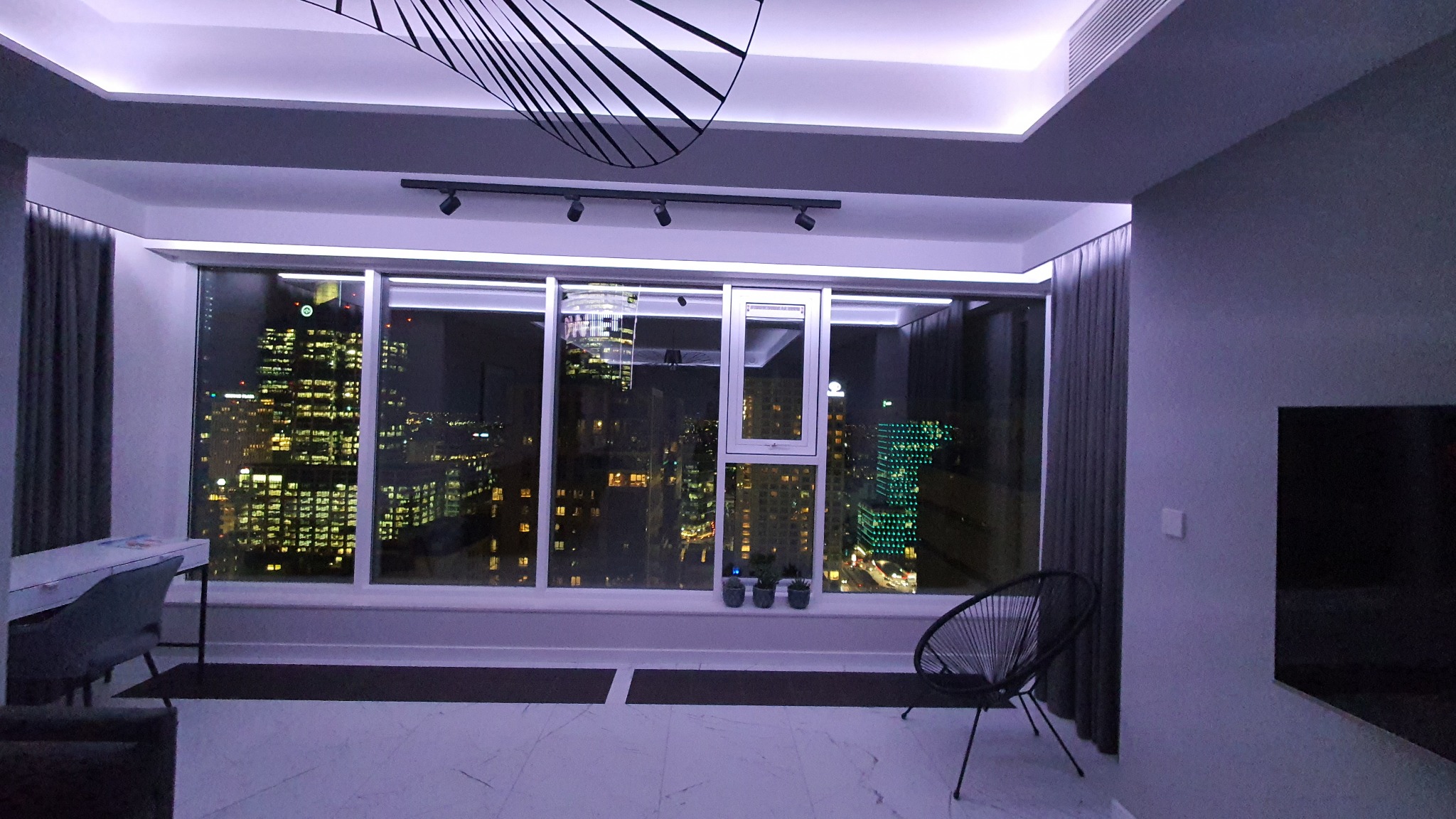 Nowoczesne wnętrze apartamentu z widokiem na nocną panoramę miasta, oświetlenie LED podkreślające sufit i okno, krzesło z czarnego drutu i biurko przy oknie.