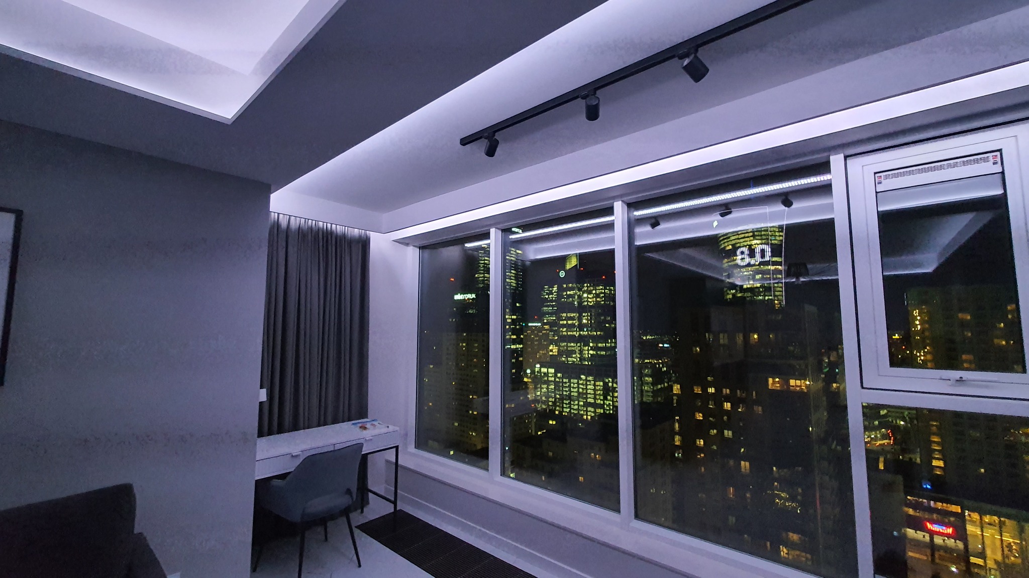 Nowoczesne wykończenie wnętrza pokoju z dużym oknem i widokiem na nocną panoramę miasta, oświetlenie LED sufitowe i punktowe, szare zasłony i białe biurko z krzesłem.