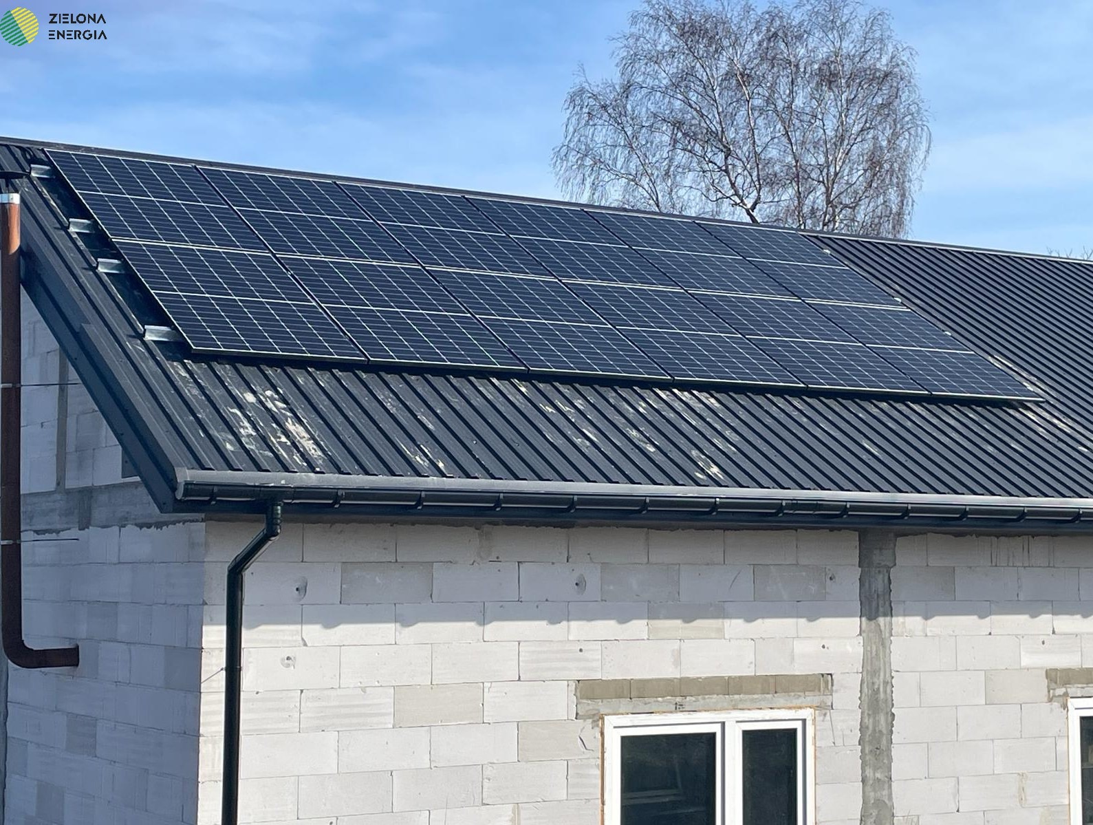 Panele słoneczne na ciemnym, metalowym dachu budynku z białego bloczka, widoczne rynny i rura spustowa, błękitne niebo w tle.