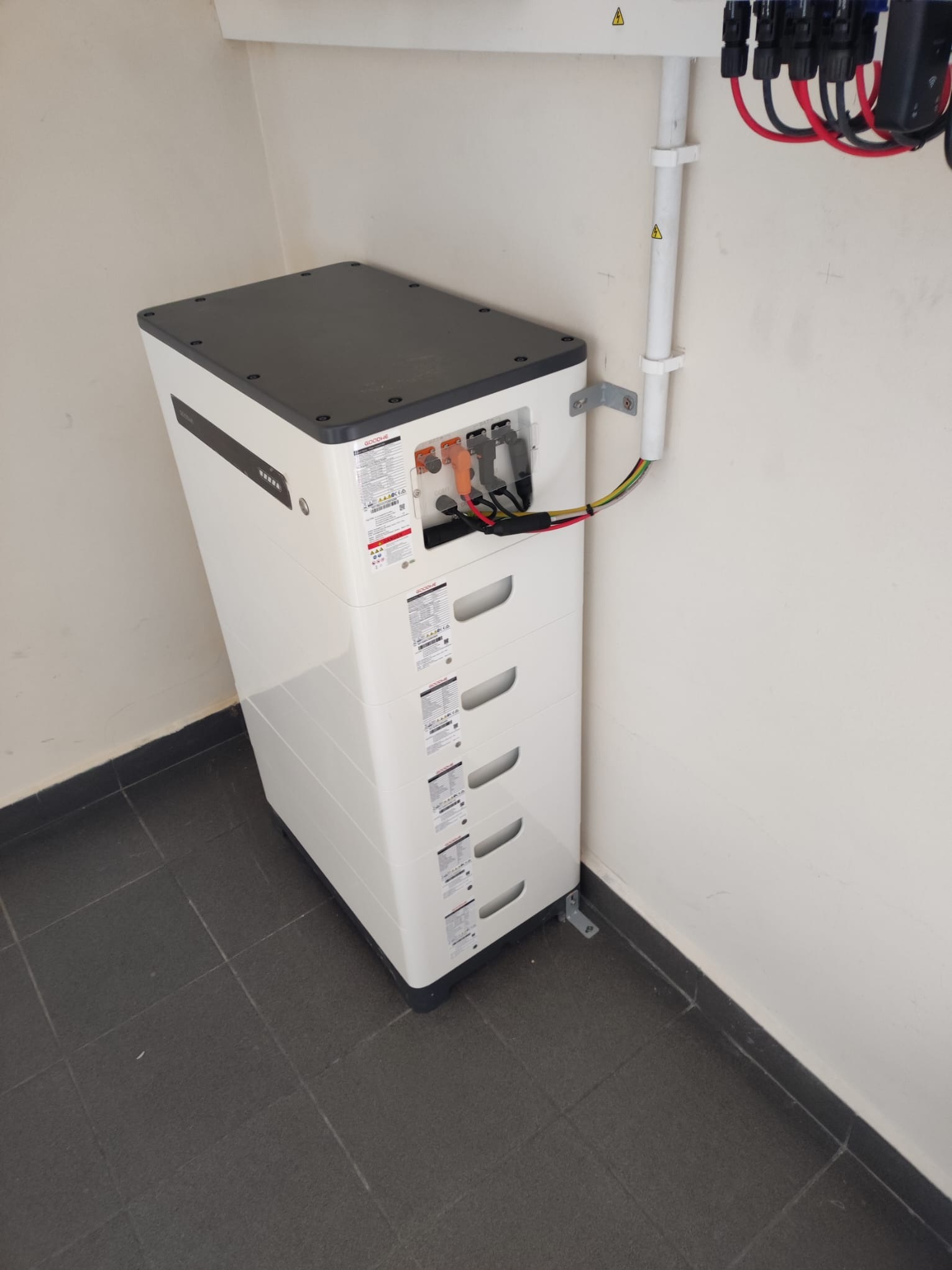 Biały, modułowy magazyn energii GoodWe zainstalowany w rogu pomieszczenia z szarą podłogą, widoczne kable i złącza, u góry rury instalacyjne z ostrzeżeniami.