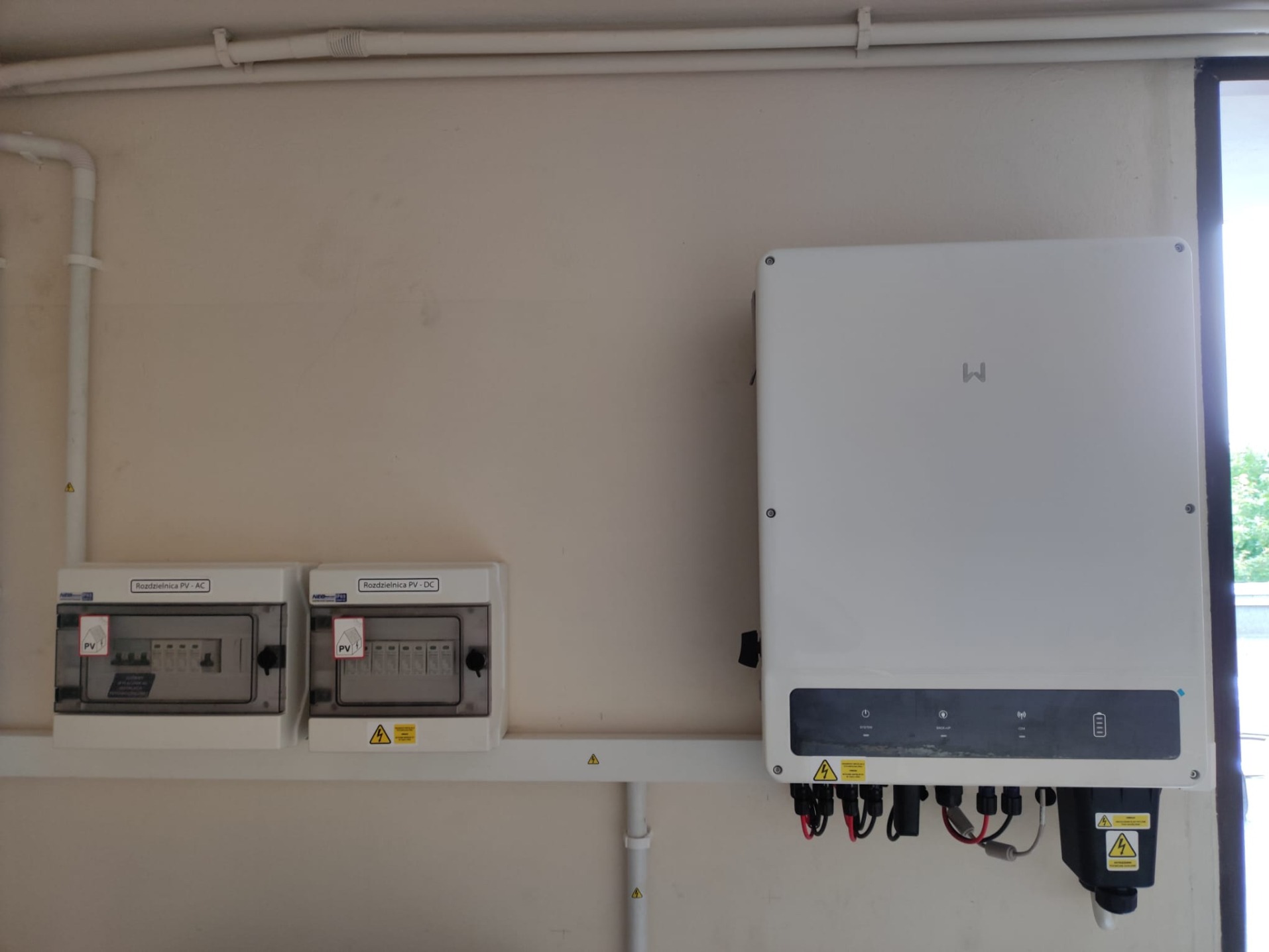 Biała obudowa falownika z podłączonymi kablami, obok dwie skrzynki rozdzielcze PV-AC i PV-DC, zamontowane na kremowej ścianie wewnątrz budynku.