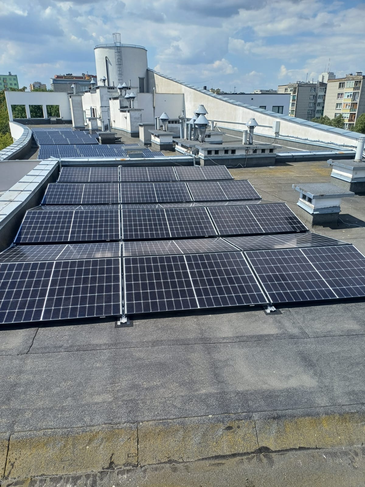 Instalacja fotowoltaiczna na płaskim dachu budynku z widocznymi panelami słonecznymi ułożonymi w rzędach, z kominami wentylacyjnymi i elementami architektury w tle, sfotografowana w jasnym...