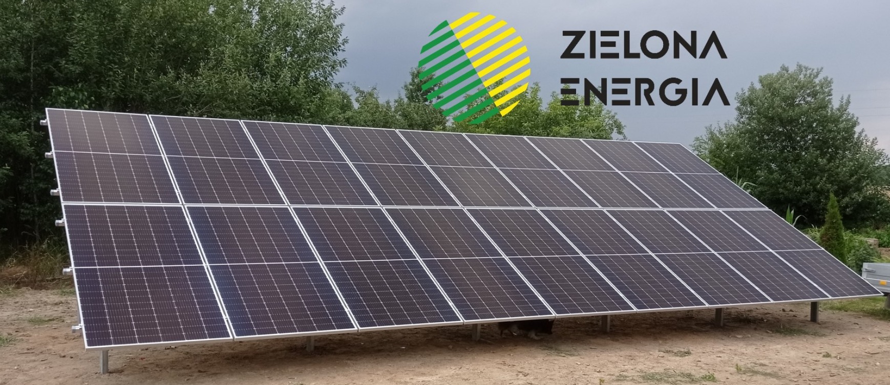 Panel słoneczny zamontowany na metalowych wspornikach, z logo firmy 'Zielona Energia' w tle, widoczne drzewa i pochmurne niebo.
