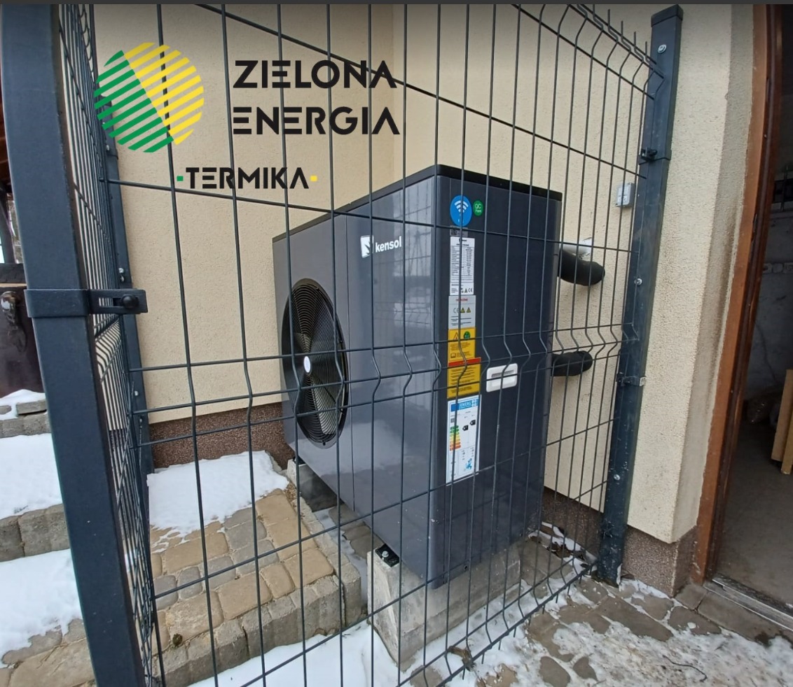 Szara pompa ciepła Kensol, umieszczona na betonowych bloczkach, za metalowym ogrodzeniem panelowym w kolorze grafitowym, z widocznym logo firmy ZIELONA ENERGIA-TERMICA w lewym górnym rogu,...