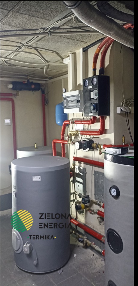 Dwa szare, cylindryczne zbiorniki akumulacyjne z logo 'Zielona Energia Termika', połączone czerwoną instalacją rurową, z widocznymi zaworami, pompami i manometrami, zamontowane w piwnicy...
