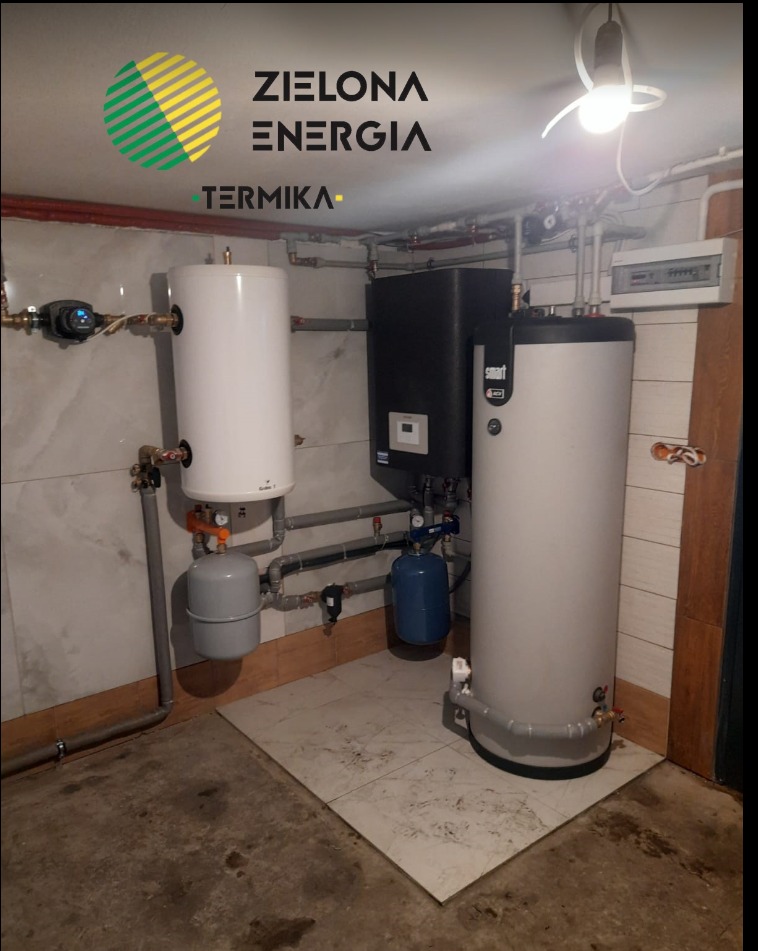 Zestaw urządzeń grzewczych w piwnicy: biały zasobnik ciepłej wody, czarny piec, szara i niebieska przepona, rury miedziane i plastikowe, logo firmy ZIELONA ENERGIA na górze.