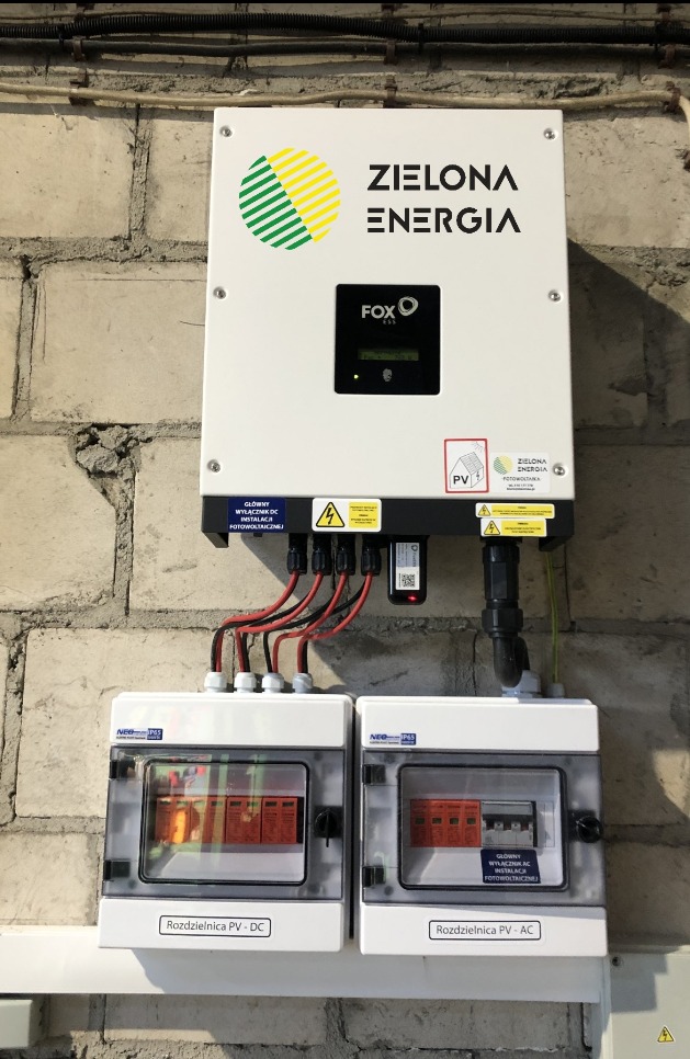 Zamontowany inwerter FOX ESS z rozdzielnicami PV-DC i PV-AC na tle ściany z cegły. Widoczne okablowanie i naklejki ostrzegawcze.