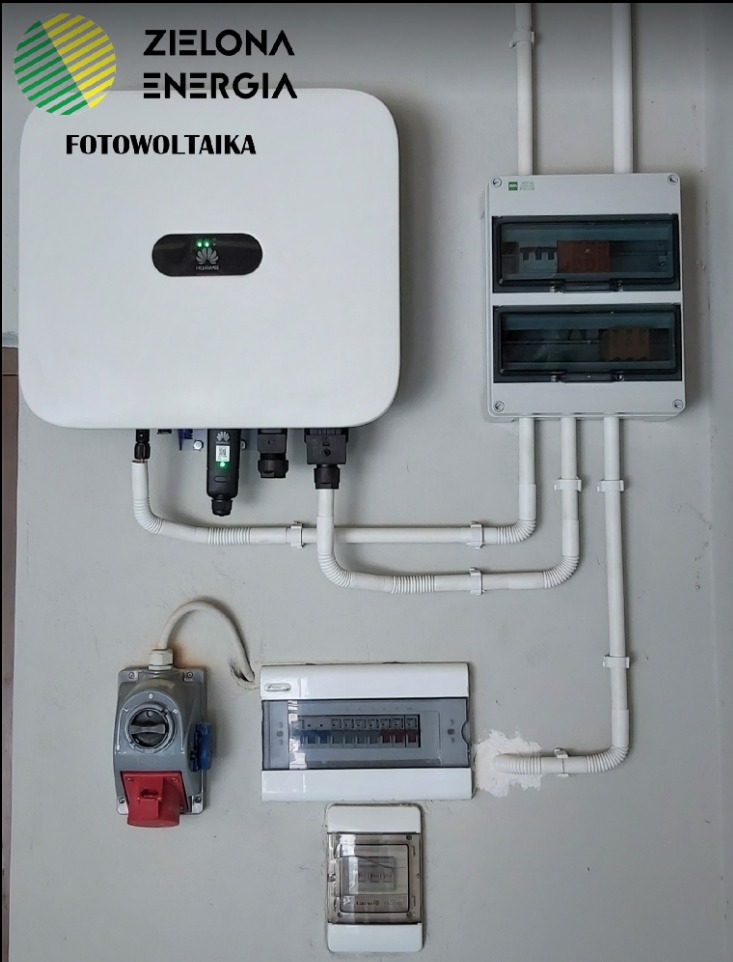 Biały falownik z logo, podłączony do instalacji elektrycznej wewnątrz budynku, widoczne zabezpieczenia i przewody w peszlach.