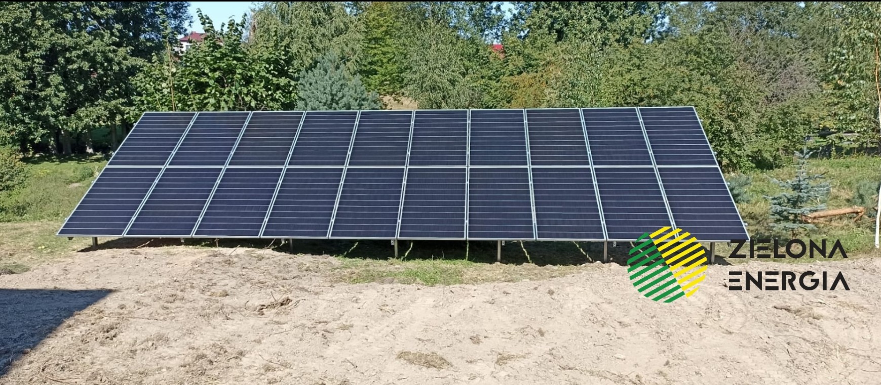 Instalacja fotowoltaiczna z logo firmy 'Zielona Energia' w prawym dolnym rogu, na tle zieleni drzew, z widocznymi elementami konstrukcyjnymi.