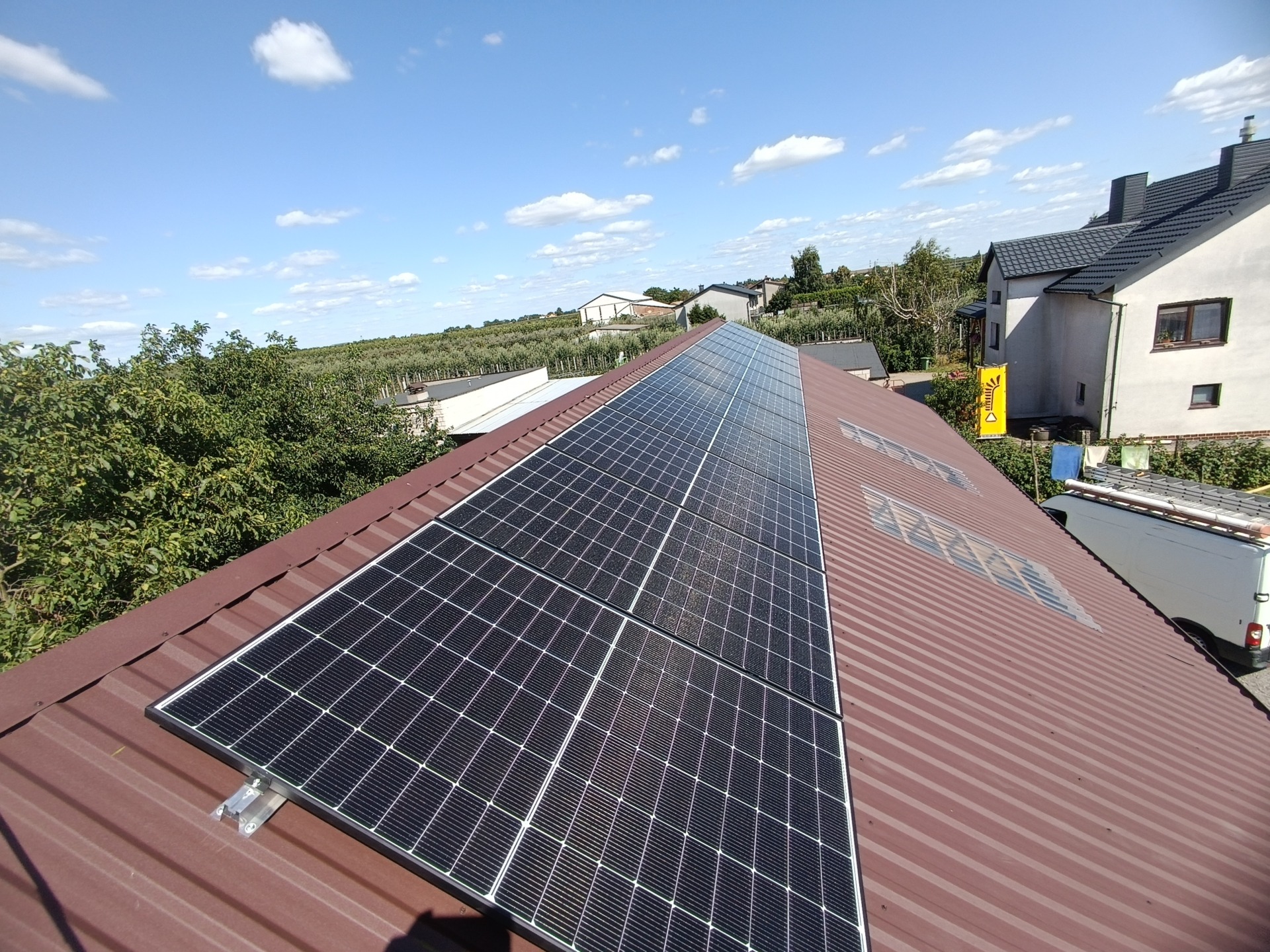 Rząd paneli słonecznych na brązowym, blaszany dachu domu jednorodzinnego, widok z góry na tle błękitnego nieba z chmurami i zielenią drzew w oddali.