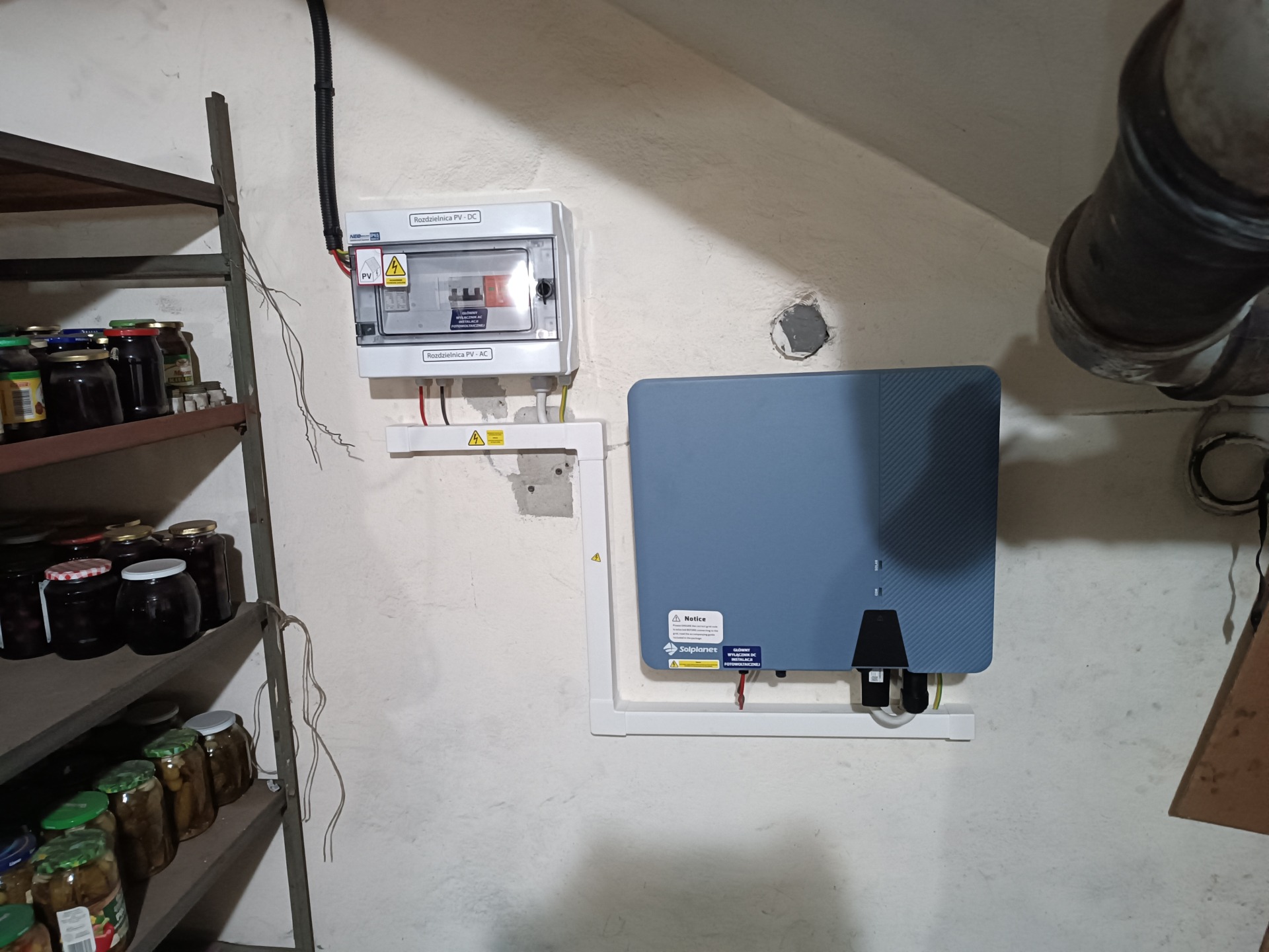 Szara skrzynka rozdzielcza PV-DC i PV-AC, niebieski akumulator energii oraz półki z przetworami w piwnicy.