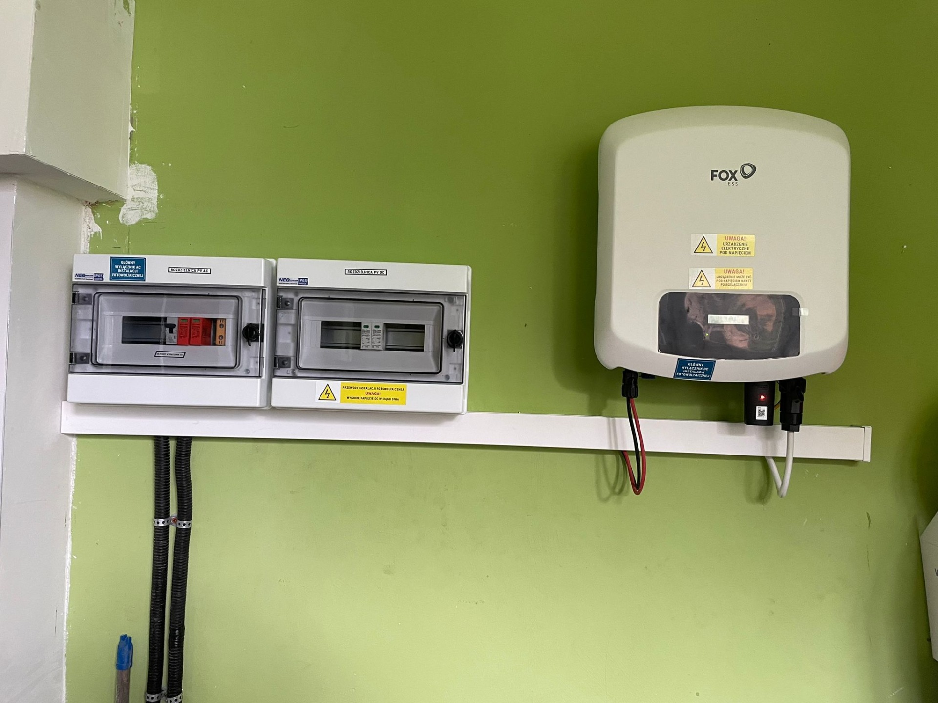 Dwa rozdzielnice elektryczne AC i DC oraz inwerter marki FOX ESS zamontowane na zielonej ścianie, przewody w peszlach. Widoczne naklejki ostrzegawcze o wysokim napięciu.