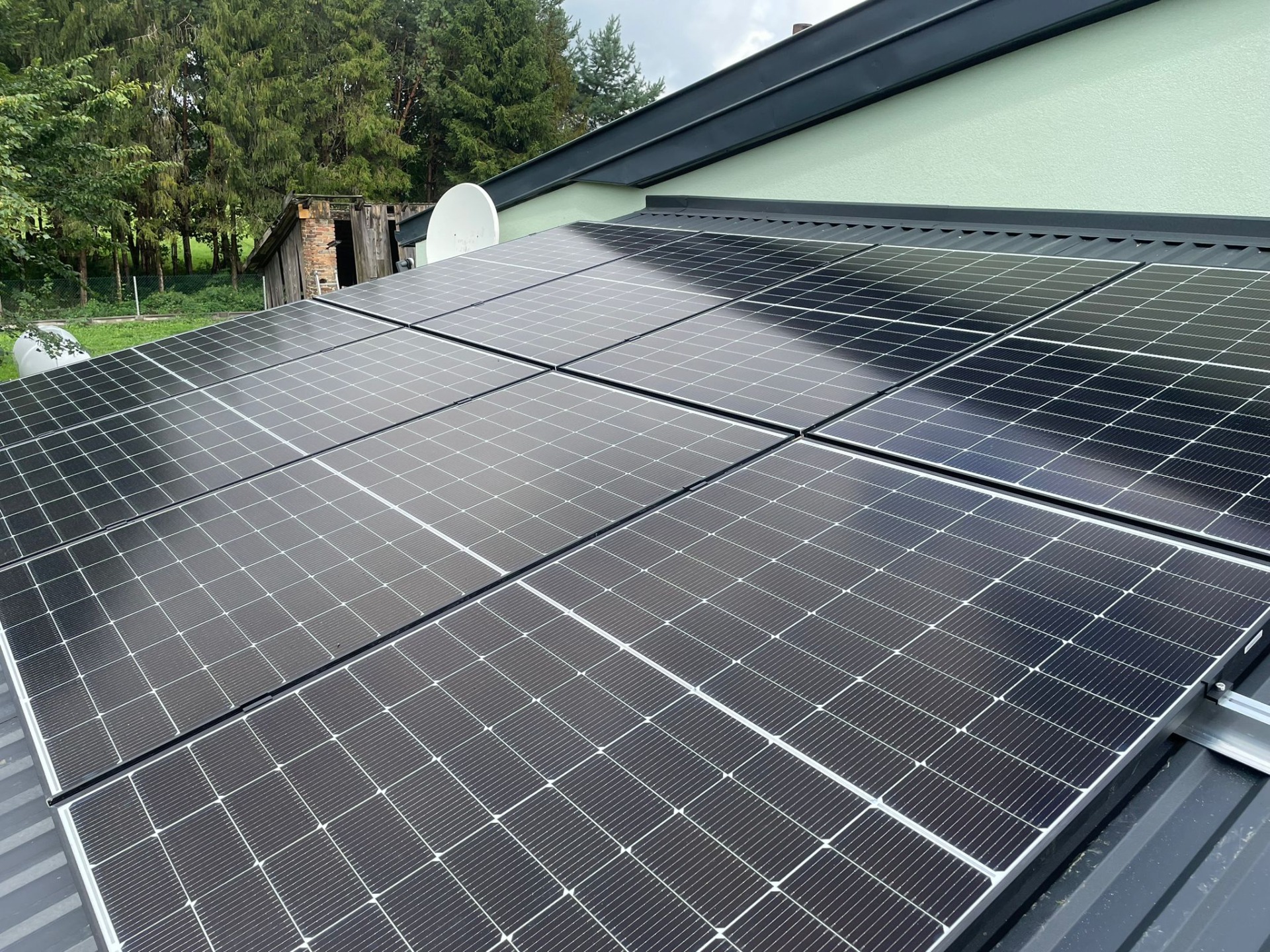 Panele fotowoltaiczne zamontowane na metalowym dachu domu jednorodzinnego, widoczne drzewa w tle i antena satelitarna.