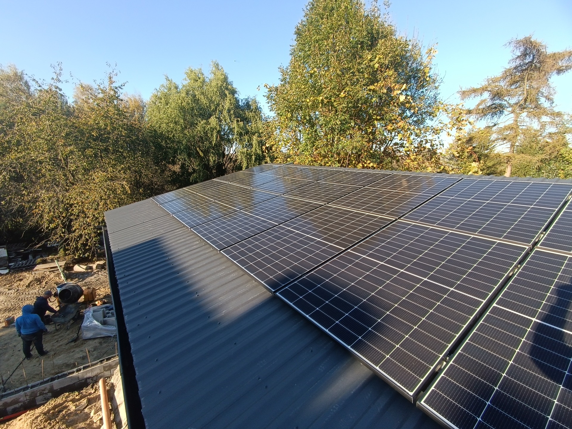 Instalacja paneli słonecznych na dachu z blachy trapezowej, widoczne drzewa w tle i pracownicy przy betoniarce.