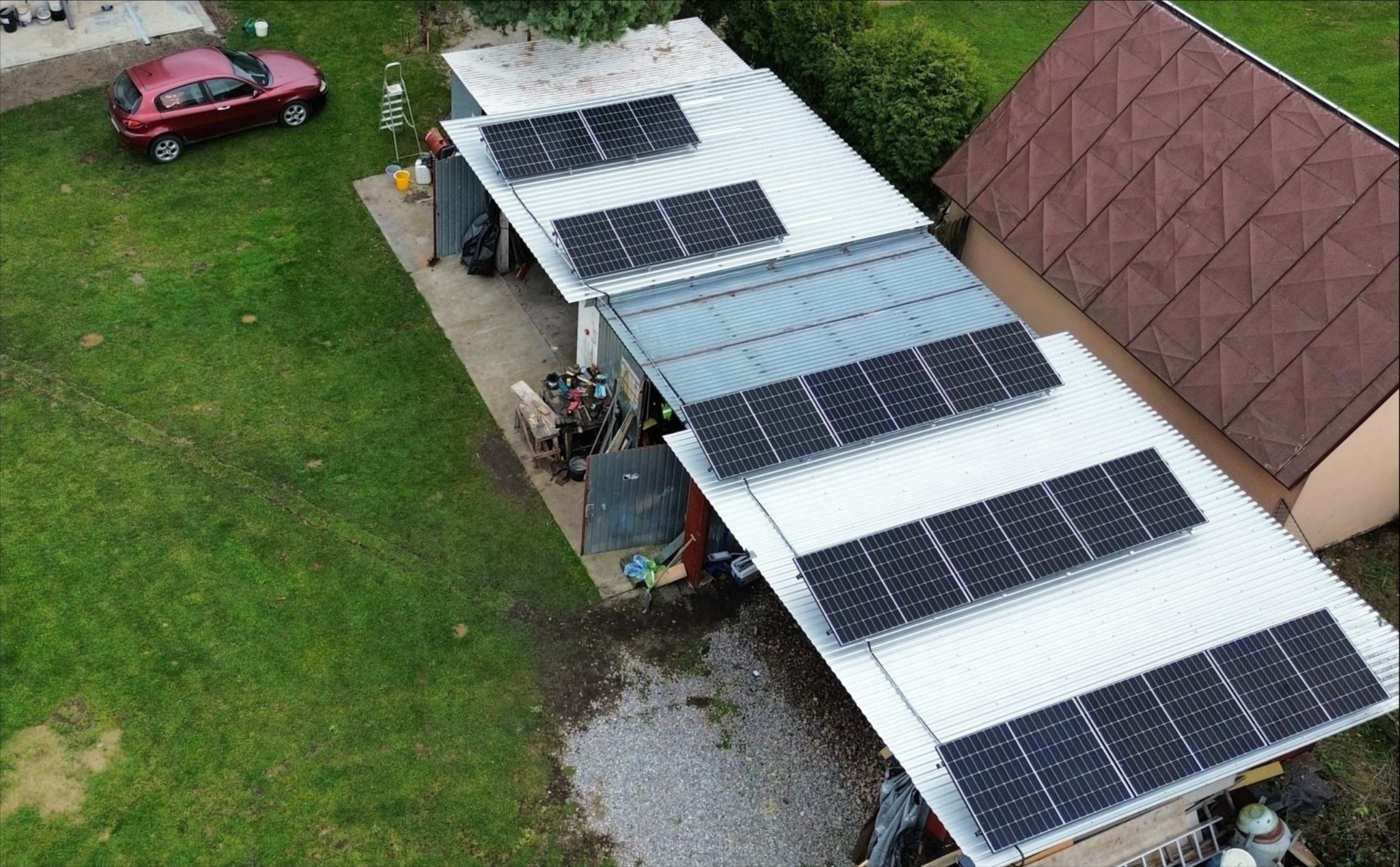 Widok z góry na budynek gospodarczy z panelami słonecznymi na dachu, obok czerwony samochód na trawniku.