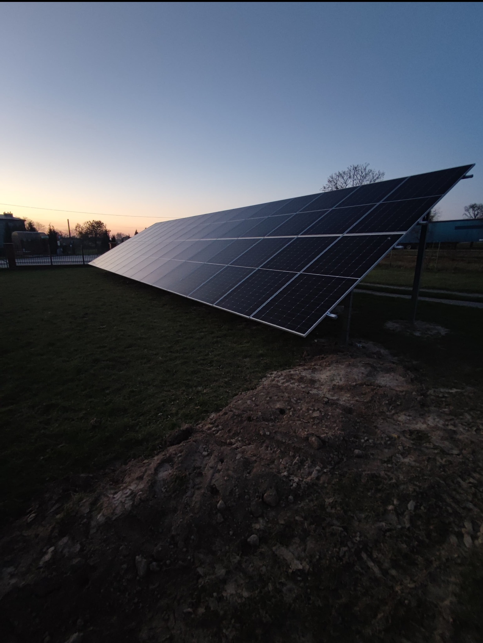 Długi rząd paneli słonecznych zamontowany na metalowych wspornikach na trawniku, sfotografowany o zmierzchu, z widocznym nasypem ziemi z przodu.