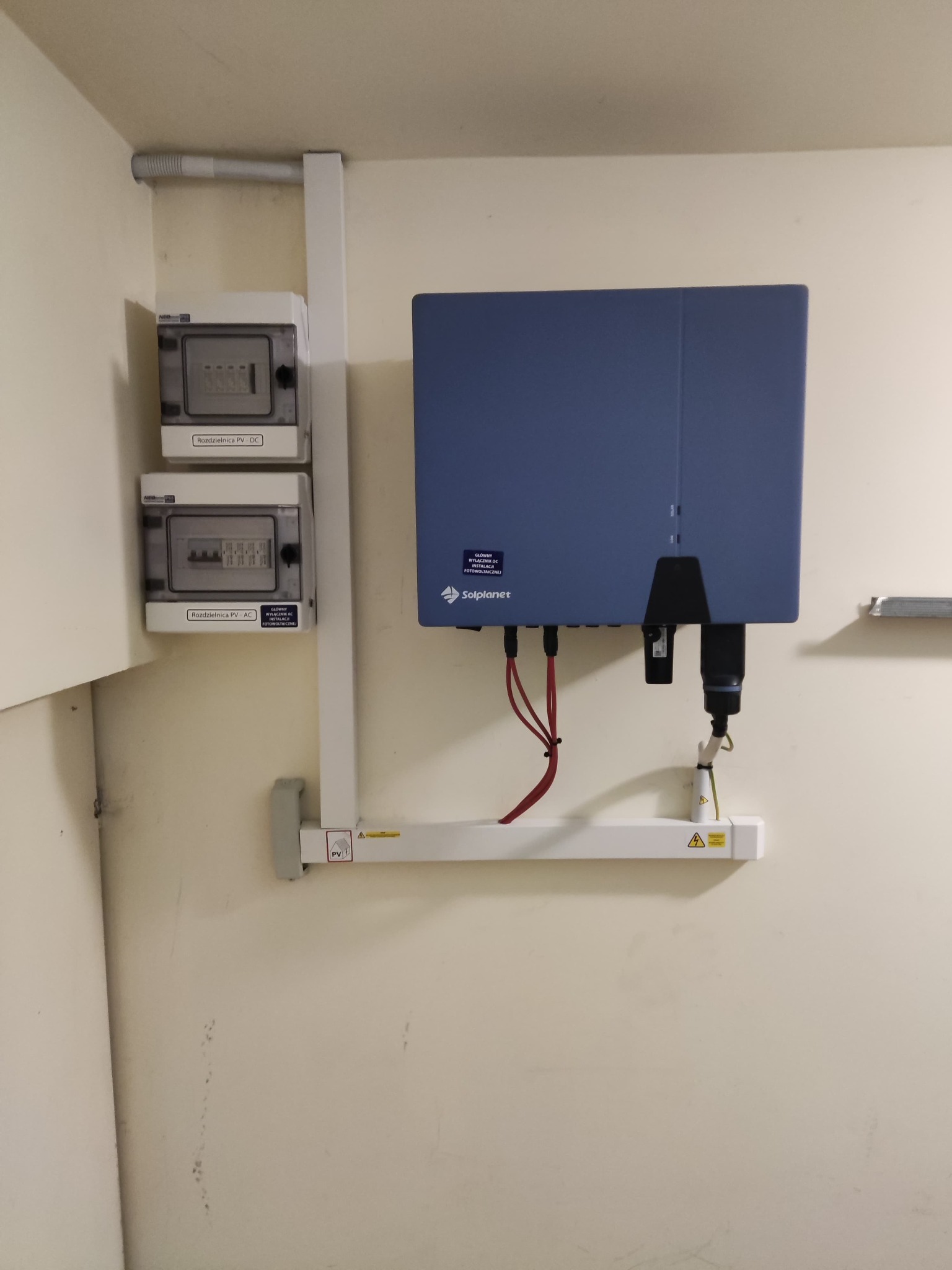 Szczegółowe ujęcie zamontowanego inwertera Solplanet w kolorze niebieskim, z widocznymi przewodami, rozdzielnicą PV DC i AC na ścianie o jasnym odcieniu, zamontowane na białych profilach.
