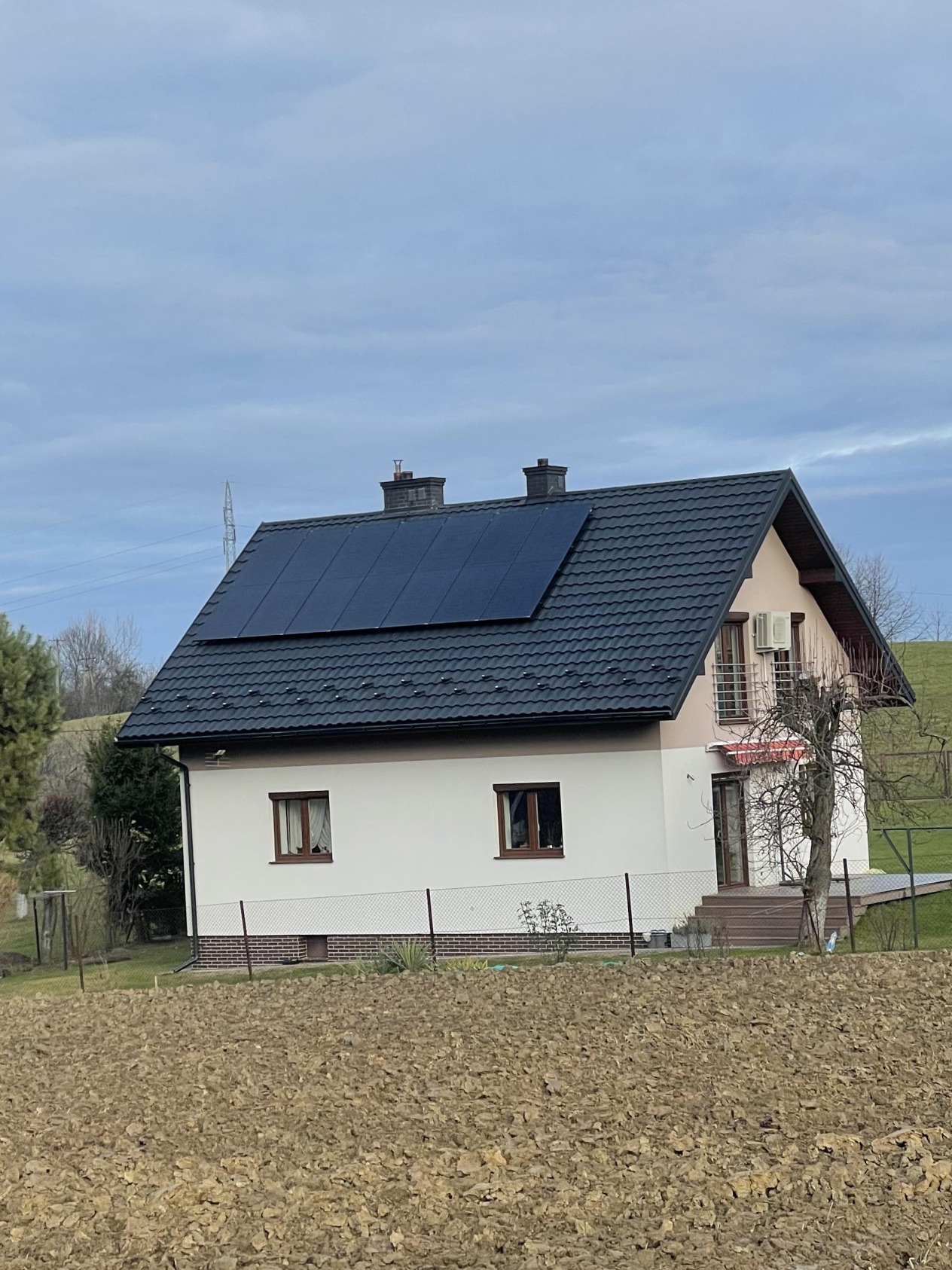 Dom jednorodzinny z ciemną dachówką i panelami słonecznymi na dachu, widoczny z pola w pochmurny dzień.