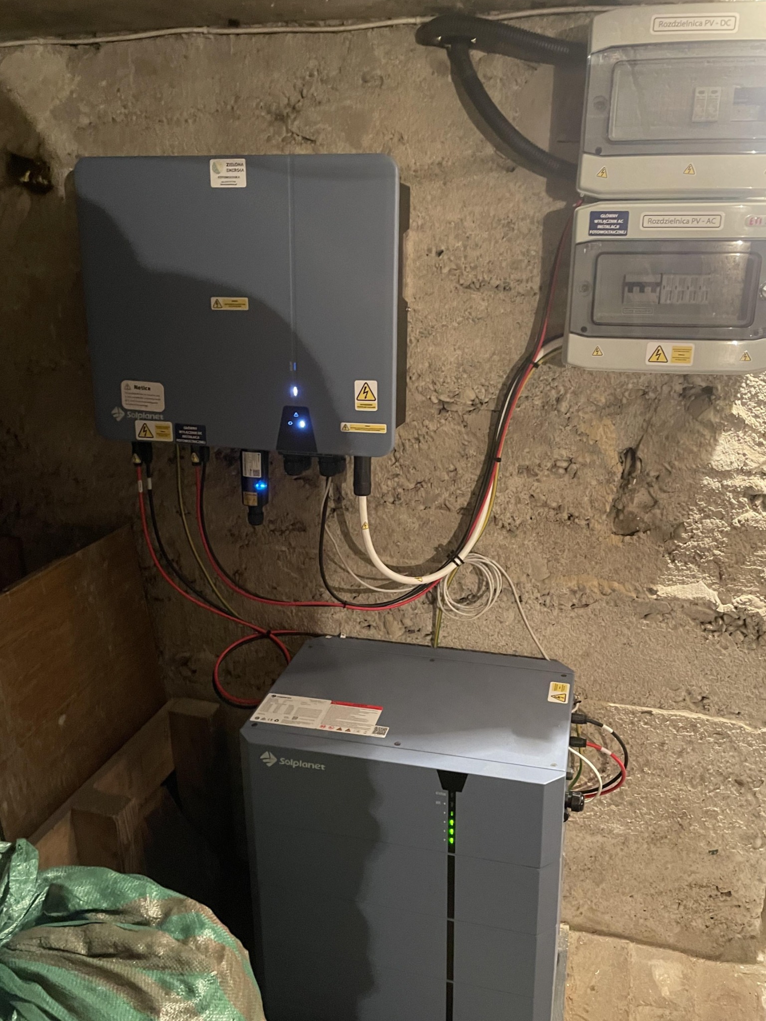 Dwa inwertery Solplanet i rozdzielnice PV-AC i PV-DC zamontowane na surowej, betonowej ścianie w piwnicy; widoczne kable i diody sygnalizacyjne.
