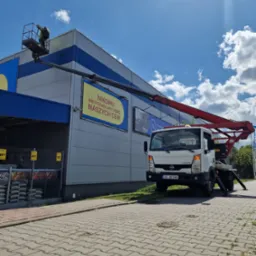 Pracownik na platformie roboczej czerwonego podnośnika koszowego, wykonuje prace na wysokości przy szarej hali z niebieskimi akcentami, widoczny baner reklamowy z hasłem 'Nikomu nie pozwolimy pobić...