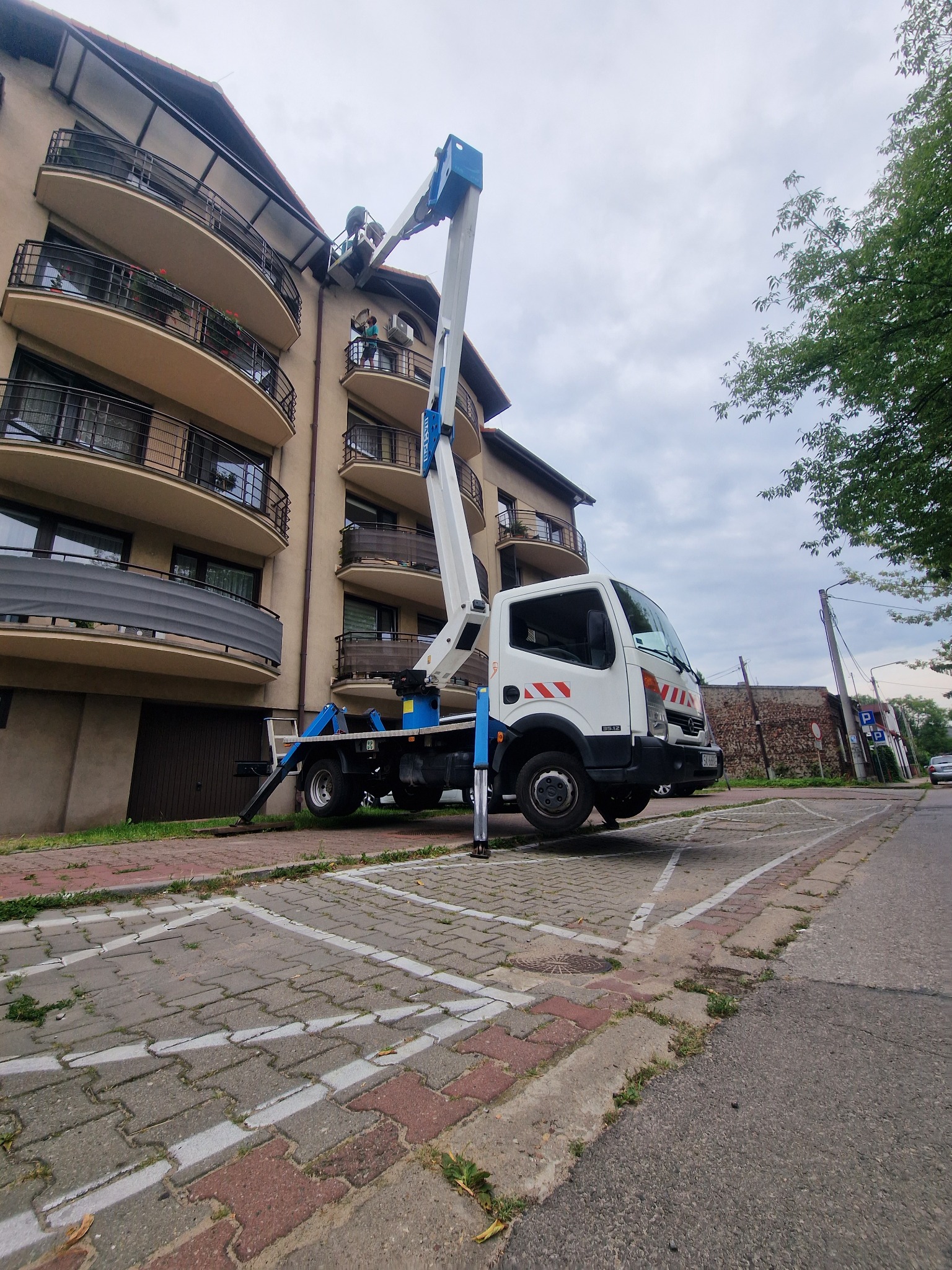 Praca na wysokości: biała ciężarówka z niebieskim wysięgnikiem na chodniku z kostki, pracownik w koszu wysięgnika przy balkonie budynku mieszkalnego.