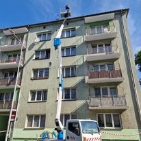 Podnośnik koszowy na tle bloku mieszkalnego z balkonami, widoczny podczas pracy na wysokości, z wysuniętym ramieniem i koszem z pracownikiem.