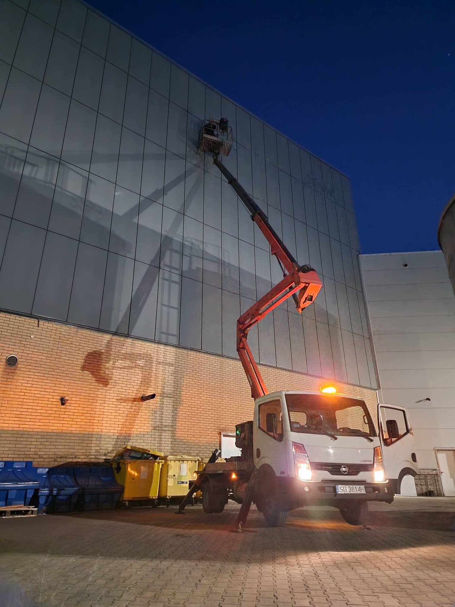 Ciężarówka z wysięgnikiem teleskopowym na tle przeszklonej ściany budynku, pracownik w koszu na wysokości, widoczne kontenery na śmieci.