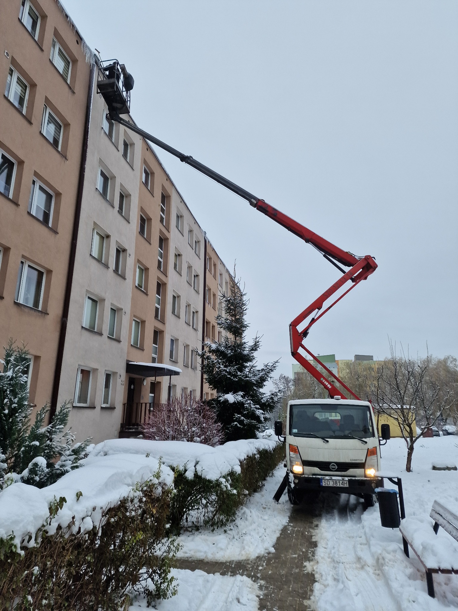 Praca na wysokościach w zimowych warunkach: czerwony wysięgnik na białym pojeździe, kosz z pracownikiem przy budynku z lodowymi sopelami, otoczenie pokryte śniegiem.