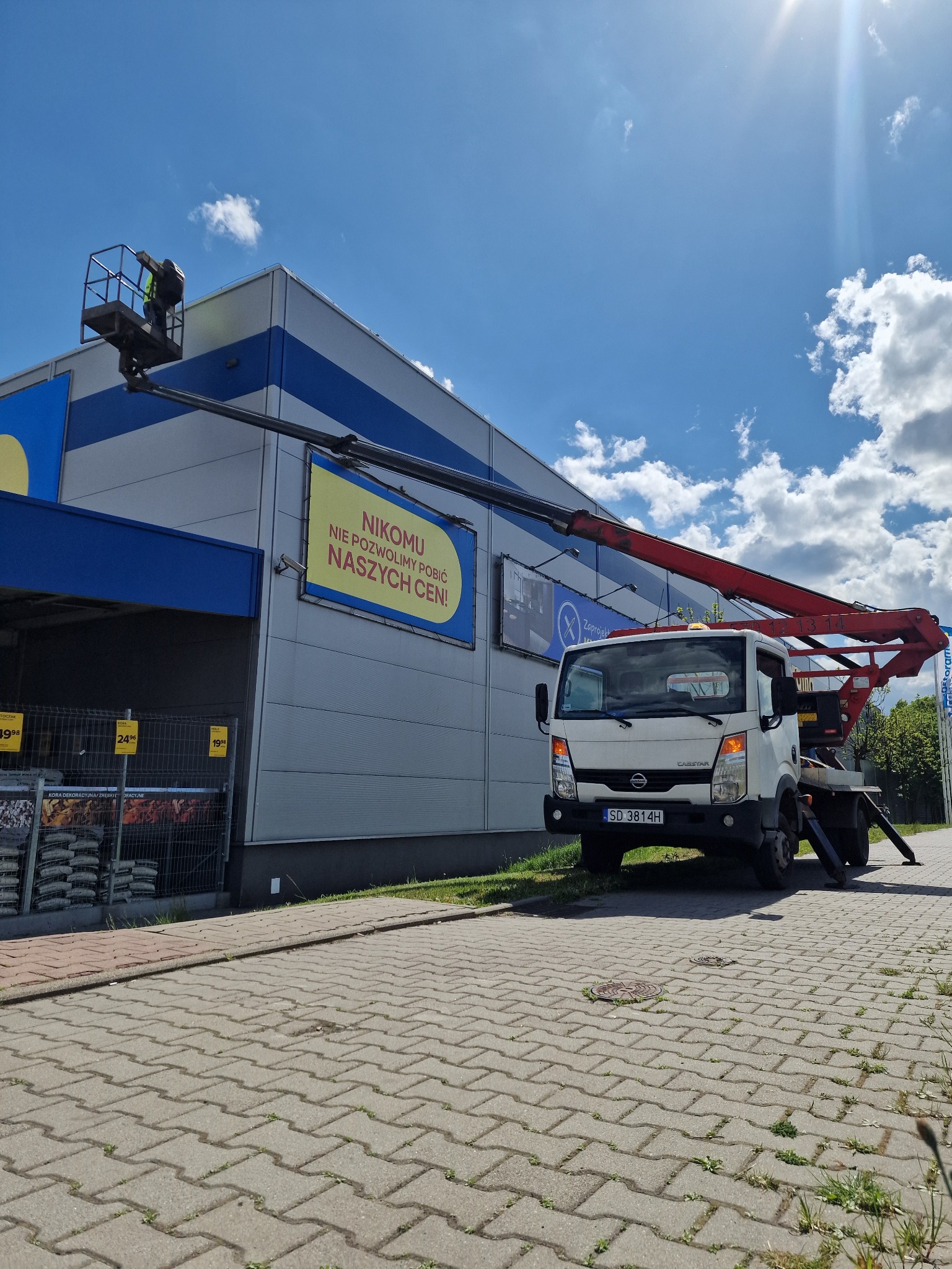 Pracownik na platformie roboczej czerwonego podnośnika koszowego, wykonuje prace na wysokości przy szarej hali z niebieskimi akcentami, widoczny baner reklamowy z hasłem 'Nikomu nie pozwolimy pobić...