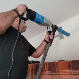 AR Klima Andrzej Banach - Mężczyzna wierci otwór w ścianie pod klimatyzator za pomocą wiertnicy z odsysaniem pyłu, widoczny uchwyt montażowy oraz fragment pomarańczowej tkaniny.